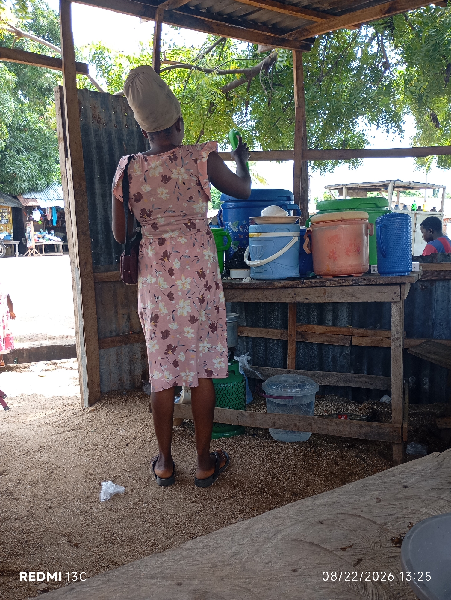 A woman wearing a light-colored headwrap and a floral print dress is standing at a wooden counter in an open-air market stall. She is holding a green bottle in her right hand. On the counter, several large plastic containers are visible, including blue, green, and orange coolers. Behind the counter, a person wearing a red and blue shirt is partially visible. The ground is covered in sand and small stones. In the background, there are trees, corrugated metal sheeting, and various market stalls. The scene appears to be in Shendam, Nigeria, based on the provided metadata.