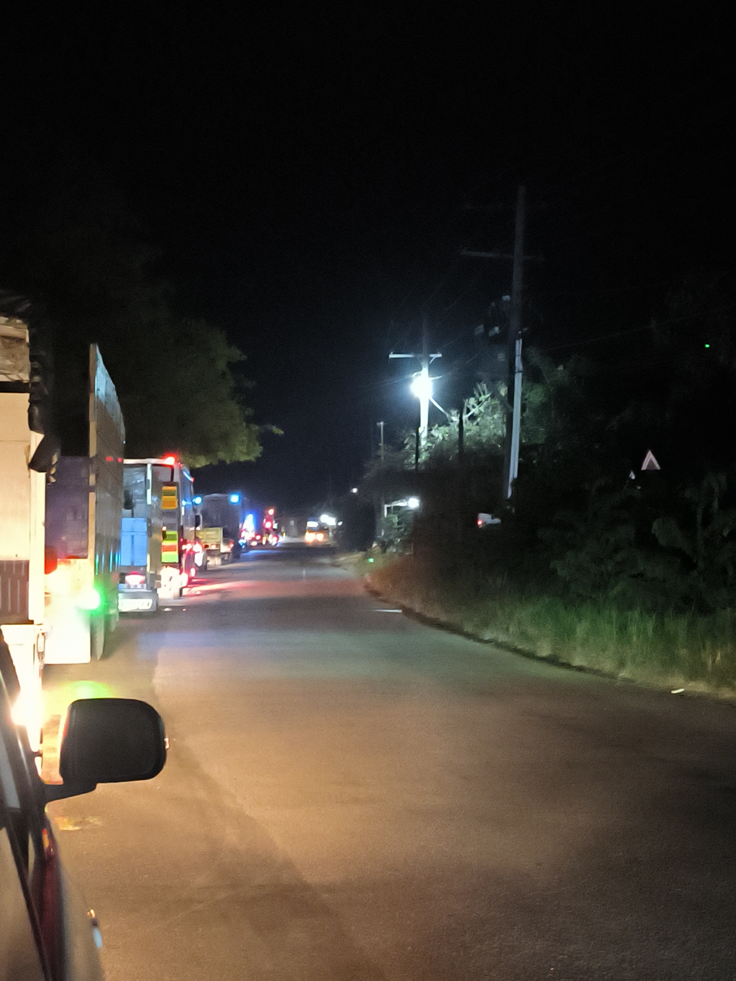 The image captures a nighttime scene on a road in Lhoknga, Indonesia. The perspective is from inside a vehicle, with a black side mirror visible in the lower-left foreground. Ahead, several large trucks are lined up on the left side of the asphalt road, their brake lights glowing red and some turn signals or hazard lights emitting green light. Various blue and white lights are also visible on the trucks and in the distance.
The road extends into the distance, appearing dark in some sections and brightly lit in others by vehicle lights and prominent streetlights mounted on utility poles. Dense vegetation, including trees and bushes, lines both sides of the road, partially illuminated by the artificial lights. A tall utility pole with power lines stands on the right side of the road, and a triangular reflective sign is barely visible among the trees further down. The dark sky confirms the time of day is night. The scene suggests vehicles are either stopped in a queue or moving slowly along the road. No visible text is present in the image.