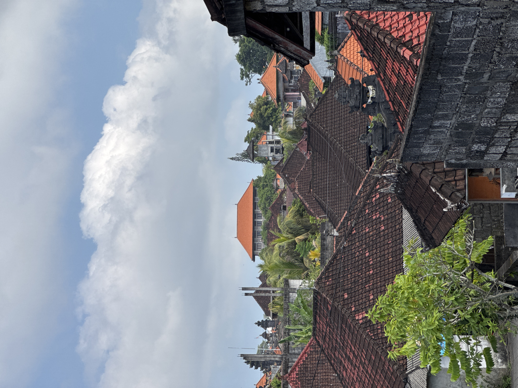 The image shows a view overlooking rooftops in Kuta Selatan, Indonesia. The rooftops are primarily made of reddish-brown tiles. A large white cloud dominates the left side of the sky, with a clear blue patch visible alongside. There is a mix of residential buildings and some taller structures visible amidst the roofs. Green foliage, including trees and tropical plants, is interspersed among the buildings. Some rooftops appear to be constructed from dark gray or black stone or brick. A green tree with thick foliage is visible in the bottom right corner of the frame.