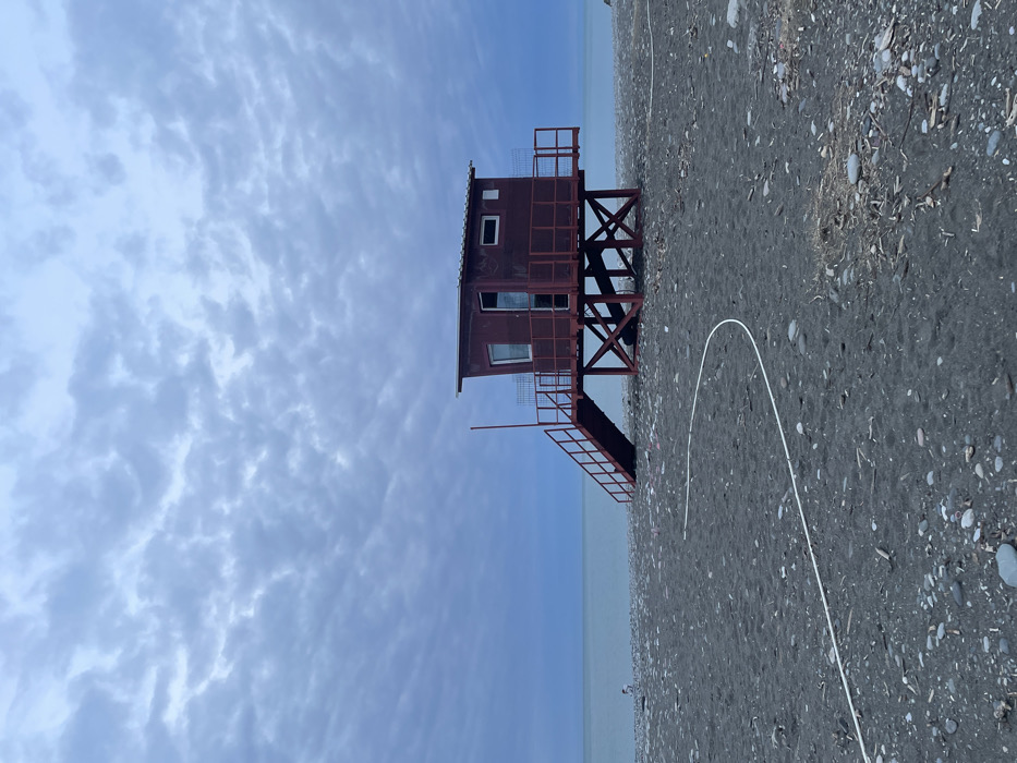 The image depicts an overcast beach scene in Batumi, Georgia. A maroon-red elevated structure, consistent with a lifeguard station or small beach hut, is centrally positioned on a dark grey pebbly beach composed of mixed sand and stones. The sky is uniformly covered with a dense layer of light blue-grey stratocumulus clouds. The calm, light blue-green sea extends to the horizon on the left.
The two-story structure features an open doorway and two windows on its visible façade, one large and one small. It is supported by an X-braced framework. A set of maroon-red stairs with handrails provides access to the elevated platform surrounding the main room. A white hose or cable lies in a gentle curve across the foreground of the dark grey beach, which is scattered with small pieces of debris. A single, indistinct human figure is visible near the waterline on the far left, appearing to be stooping or crouching. No other individuals or significant actions are observable.