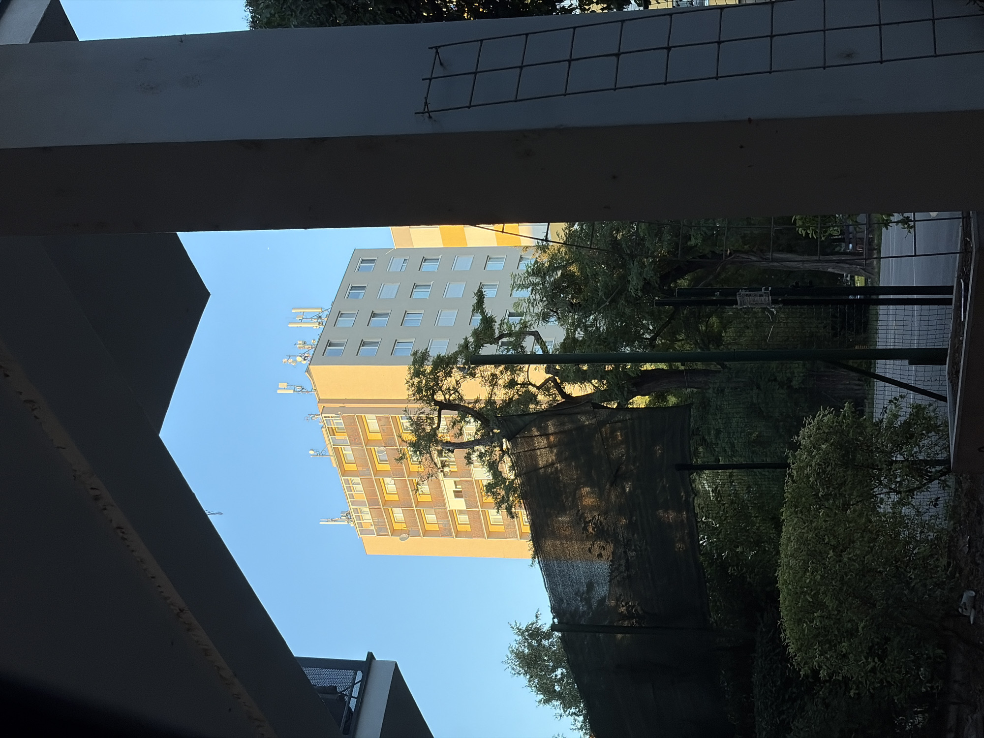 The image shows a tall residential building with a yellow facade and balconies, seen through an overhang. The building has several antenna arrays on its roof. In the foreground, there are trees and a black net stretched between poles, partially obscuring the view. The scene appears to be from a residential area in the city of Nitra, Slovakia. The sky is clear and blue. The image has a clear perspective, indicating it was taken from an elevated position, likely a balcony or window.