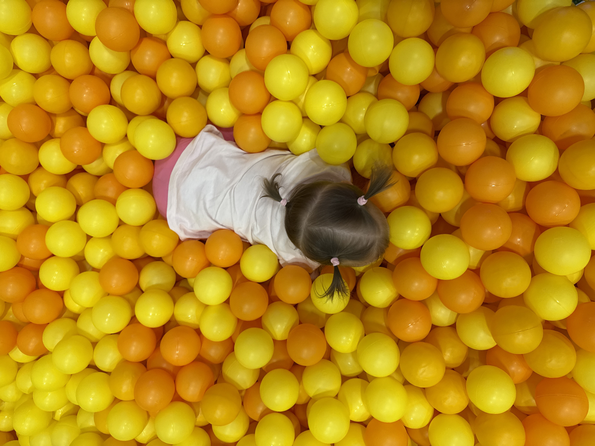 A high-angle, full shot depicts a child lying in a ball pit filled with yellow and orange plastic balls. The child is wearing a white top and pink bottoms. The back of the child's head and two pigtails are visible. The location is Kommunarka, Russia.