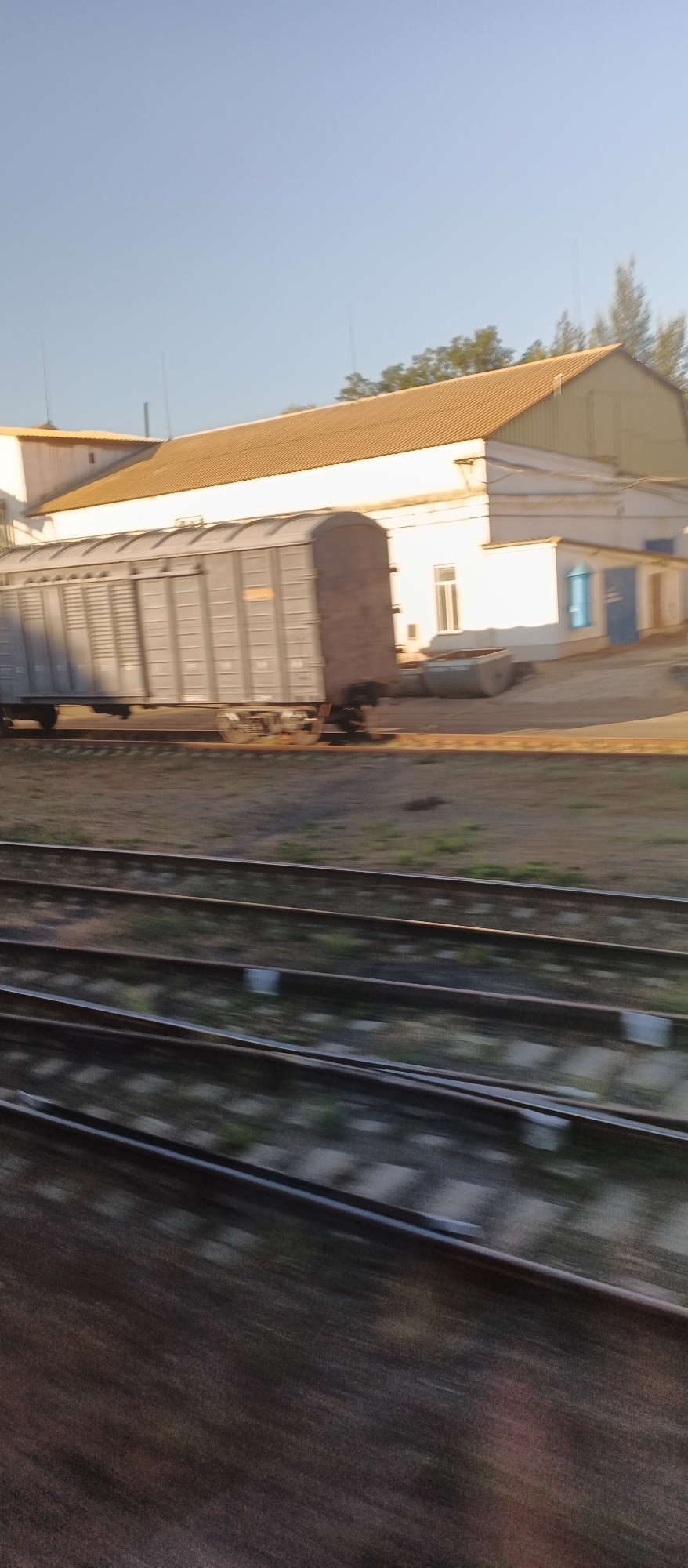 An outdoor, eye-level, slightly blurry, medium shot shows a train car and buildings in Khlystunivka, Ukraine. The train car is a dark grey freight car with ridged sides and is positioned horizontally across the middle of the frame. The car is on train tracks, with several sets of parallel tracks visible in the foreground, running horizontally. Behind the train, a light-colored building with a brown roof extends across the frame. The building has several windows and doors, one of which is bright blue. Trees are visible behind the building, and the sky is a clear, pale blue. The scene appears to be in daylight, possibly late afternoon, given the shadows and lighting. There are no people visible in the image.