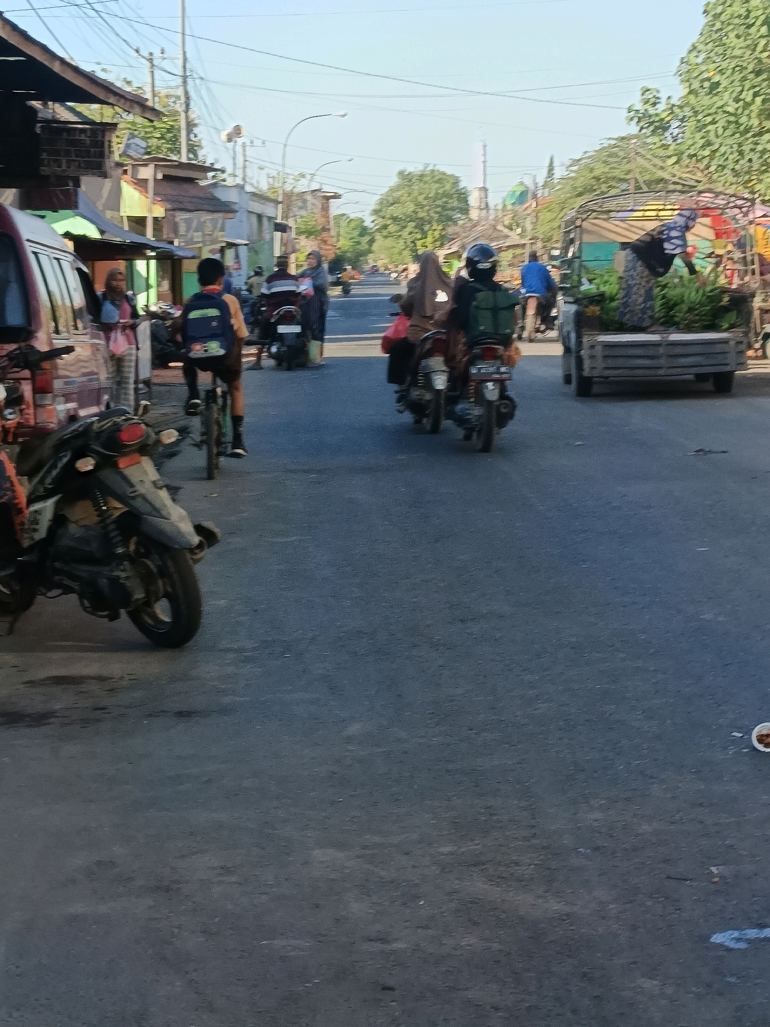 The image depicts a street scene in Blega, Indonesia, likely during daytime. The foreground features a dark-colored motorcycle. In the mid-ground, several people on motorcycles are visible. One motorcycle carries two individuals, and another has a rider. Further down the street, a truck is laden with what appear to be bananas. The surroundings include a row of shops or market stalls along the left side of the road, with power lines and poles overhead. Trees are present on the right side of the street. The road surface is asphalt, with some debris and a discarded cup visible. The sky is clear and blue. The overall impression is of a busy local street in a Southeast Asian town.