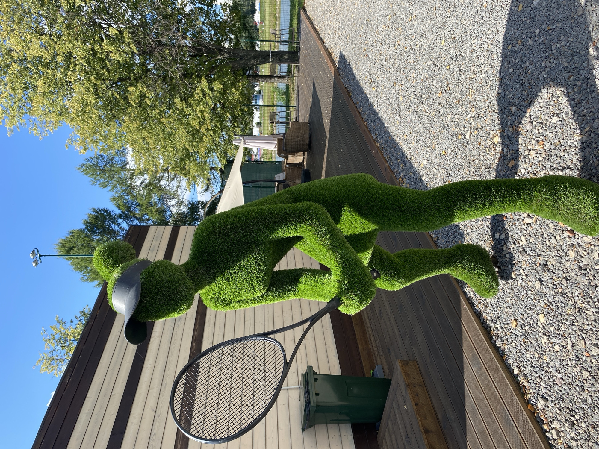 An outdoor scene features a topiary figure shaped like a person playing tennis, complete with a tennis racket and a black visor. The figure is made of green grass and stands on a gravel path. Behind the figure, a wooden deck is visible, with a wooden bench and a green trash bin. In the background, there is a building with horizontal wooden siding, trees, and a body of water. The sky is blue with scattered clouds, and sunlight casts shadows on the ground.
