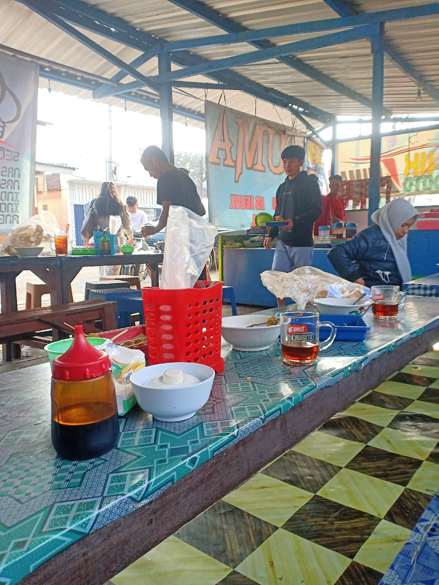 The image depicts an outdoor food stall in Jatisrono, Indonesia, under a blue metal roof supported by metal beams. Sunlight streams in from the right, casting shadows and creating a bright atmosphere. Several people are visible: a man in a black t-shirt is serving food from behind a counter, a young man in a black hoodie is holding a plate of food, a woman in a blue jacket and grey hijab is seated at a table, and another person is partially visible behind the serving counter.
The foreground features a table covered with a patterned tablecloth. On the table are several items, including a red plastic basket filled with plastic bags, a white bowl with a lid, a bottle of soy sauce with a red cap, and two clear mugs filled with a dark liquid, likely tea. In the background, there are banners with text, likely advertising food items, and a counter area where food is prepared and served. The overall environment suggests a casual dining setting, possibly a local eatery or warung.