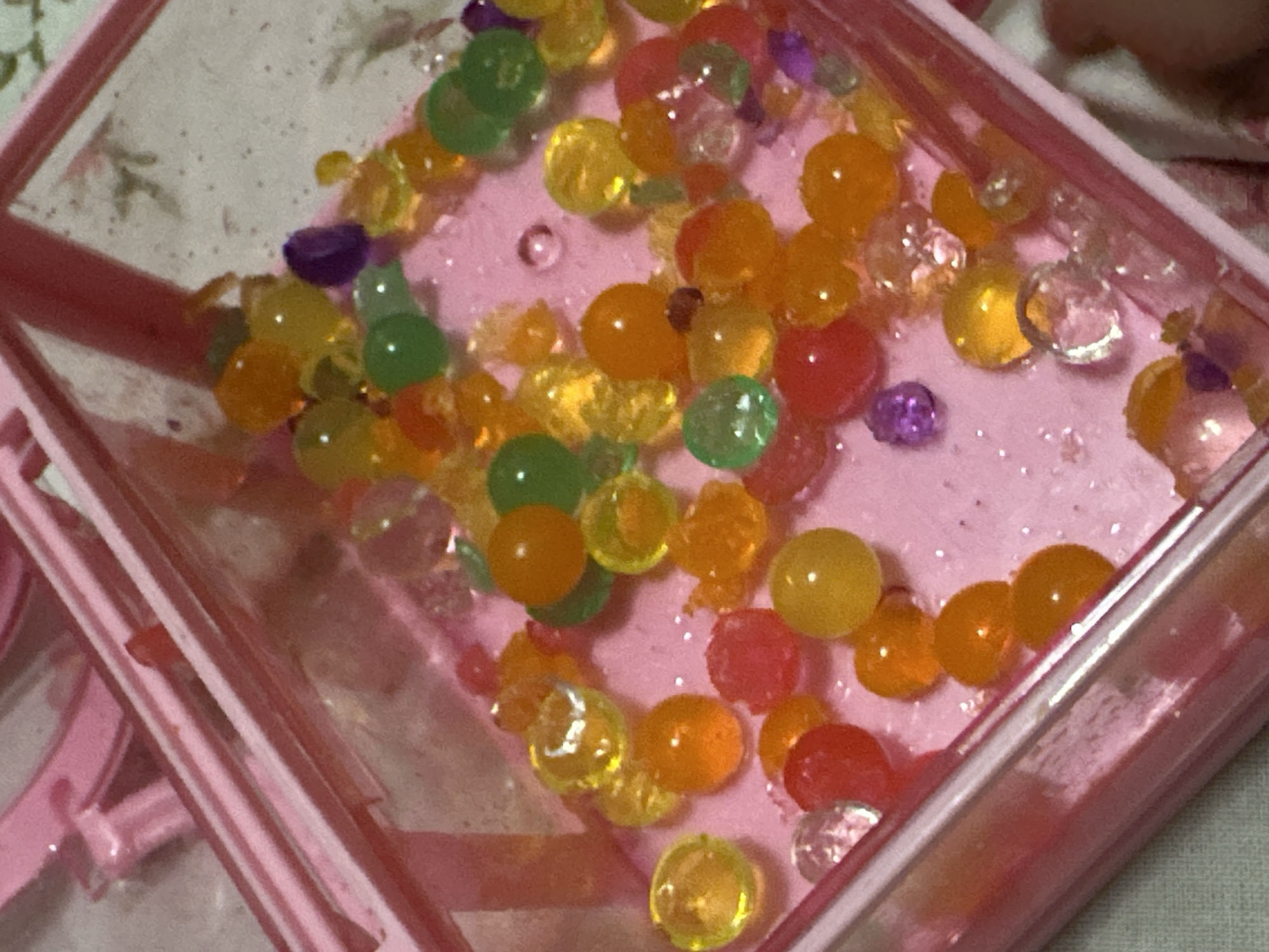 A close-up shot of a pink plastic container filled with an assortment of colorful water beads of various sizes. The beads are primarily translucent shades of green, yellow, orange, red, and clear, scattered randomly across a light pink base. The pink container is angled slightly, showing the edge and corner details of its structure. There is an absence of people in the scene, and no actions or interactions are taking place. The environment is limited to the contents of the container and the immediate surrounding area, which appears to be a flat surface. The image does not provide any geographic context related to Uva, Russia.