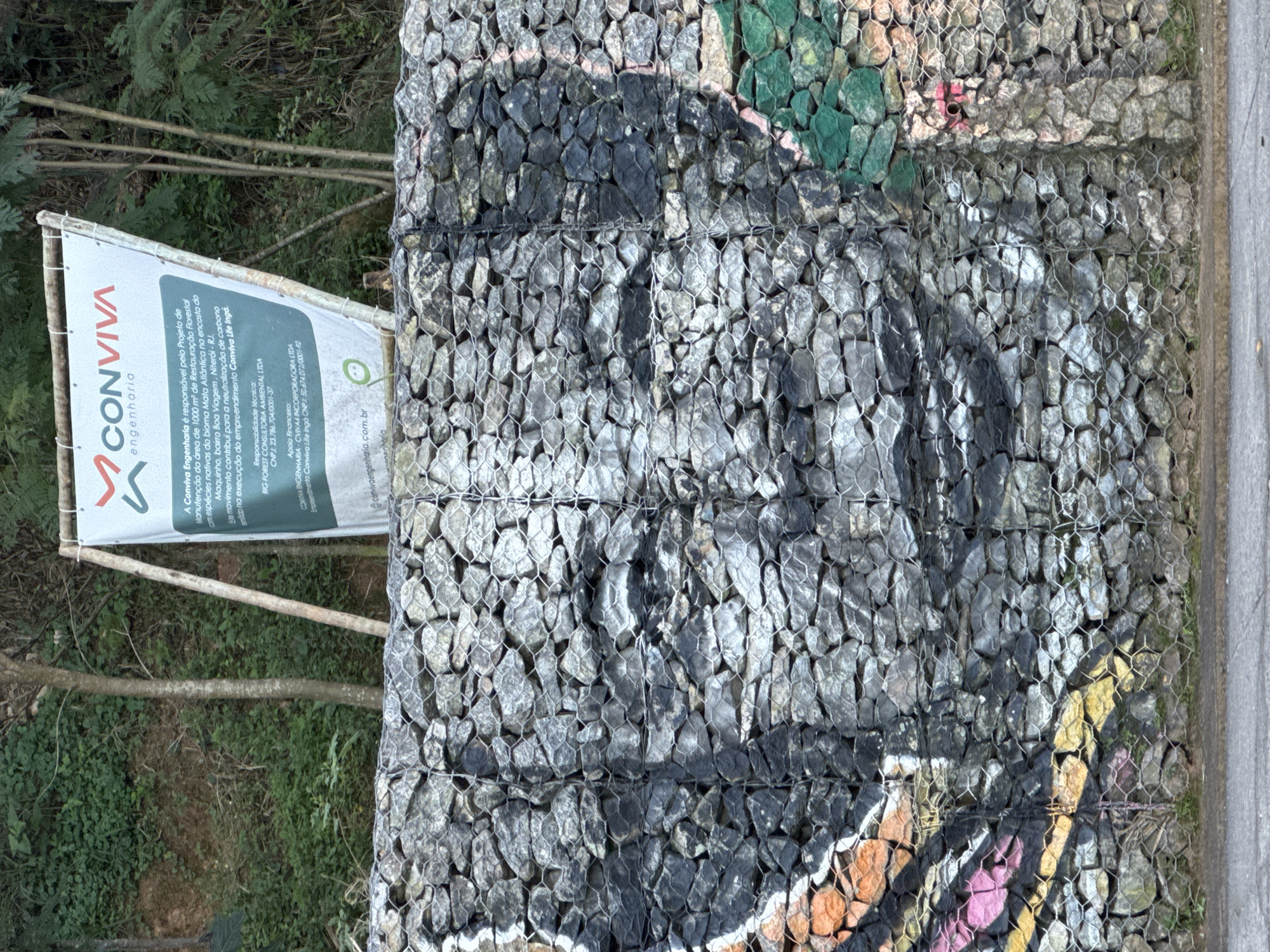 The image depicts a gabion wall covered with graffiti, featuring a mural of a face painted in shades of black and gray. To the left of the mural, there are abstract graffiti elements in orange, yellow, and pink. A white banner with the CONVIVA engenharia logo and text is visible at the top of the image, supported by wooden poles. The banner details a forest restoration project in the Macarenko, Bairro Boa Viagem neighborhood of Niterói, Rio de Janeiro, Brazil. The background consists of a densely vegetated hillside with trees. The lower part of the image shows a concrete surface, likely a sidewalk or road, in front of the gabion wall.