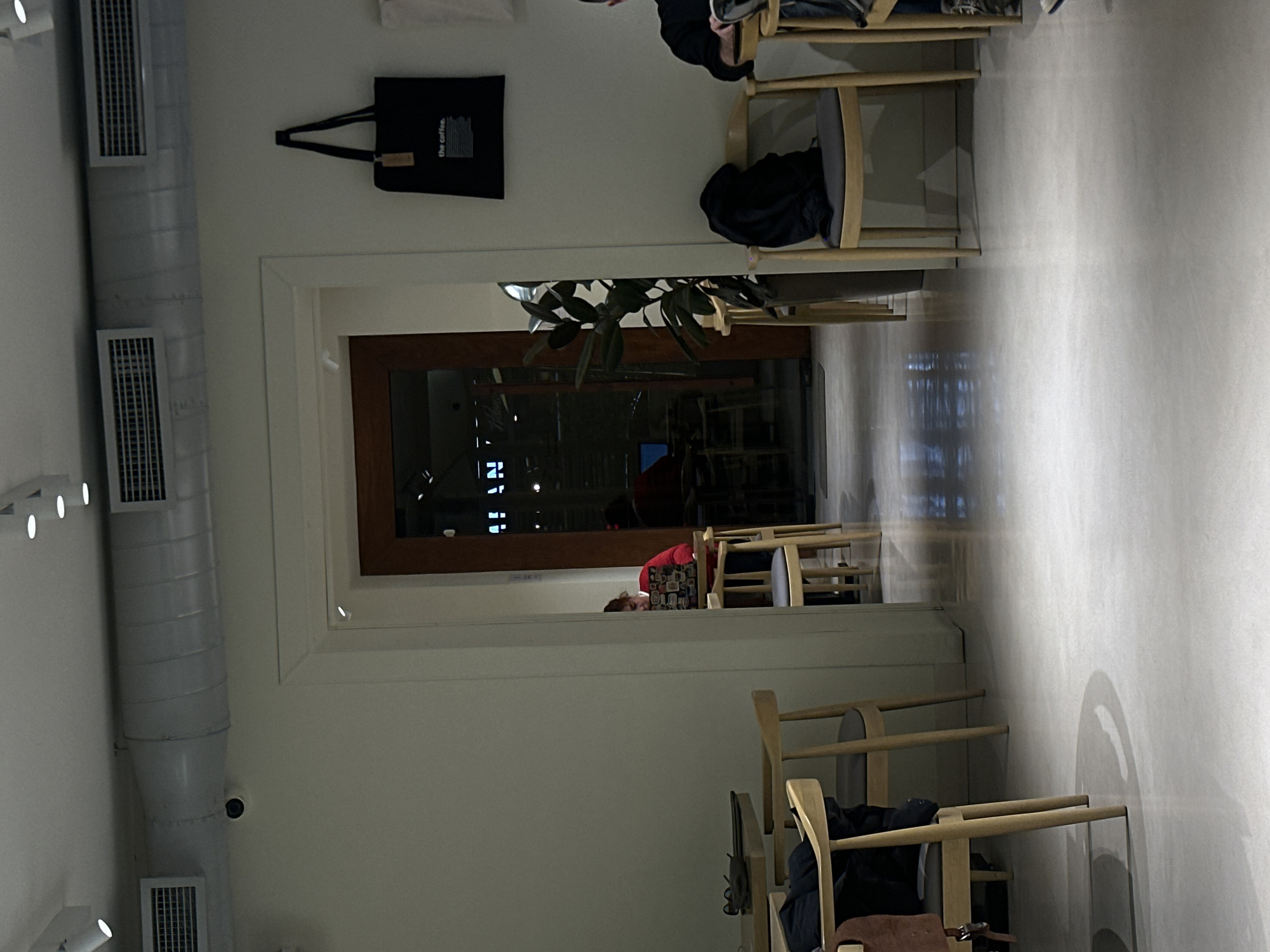 An indoor scene in Yerevan, Armenia shows a dining area with light-colored walls, wooden chairs, and a doorway leading to another room. A person with red hair and wearing a red patterned shirt is seated at one of the tables. The walls are adorned with items like a black tote bag and what appear to be ventilation ducts. The overall lighting is soft, creating a calm atmosphere.