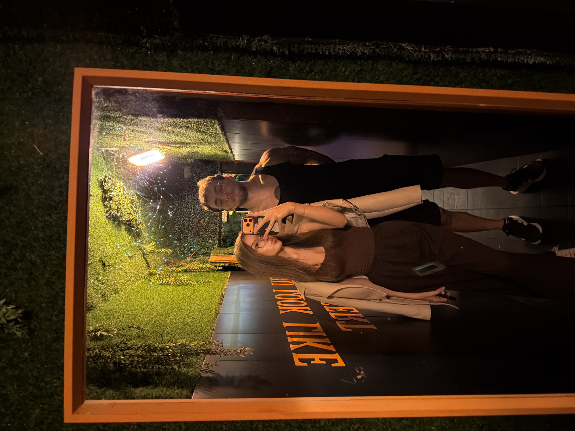 A vertical, medium shot captures a couple taking a mirror selfie in a dimly lit establishment adorned with artificial greenery and orange lighting. The woman, positioned closer to the mirror, is in the foreground, her body angled towards the camera. She has long, brown hair and wears glasses, a brown camisole, and dark trousers, with a light-colored jacket draped over her shoulders. She holds a smartphone in her right hand, capturing the reflection. To her left, a man stands slightly behind her, wearing a black tank top and dark shorts. He is looking into the mirror with a neutral expression. The background of the mirror reflection shows a textured green wall with hanging artificial plants and some string lights, suggesting an indoor space with a naturalistic theme. The mirror itself has a thick, orange frame. On the black wall behind the couple, large orange letters are visible, forming words that appear to be a quote or phrase. The overall impression is a candid moment captured during an outing.
