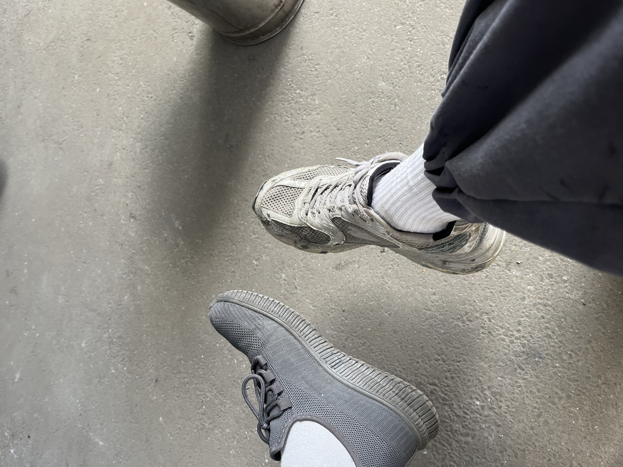 The image shows a close-up, top-down view of two pairs of feet wearing athletic shoes and socks, resting on a concrete surface. The feet are positioned on the left and right sides of the frame, with a noticeable gap between them. The shoe on the left is a dark gray, low-top sneaker with laces. It appears to be relatively clean. The shoe on the right is a lighter gray and appears to be well-worn and possibly dirty. The individual on the right is wearing white socks and dark pants. The concrete surface is a neutral gray color, with some texture and small debris visible. The lighting suggests an outdoor setting, possibly under a shadow. No people are fully visible, only their lower extremities. There is a cylindrical object in the upper right corner of the frame, partially out of focus, which could be a trash can or a post. The scene is devoid of any specific identifying markers of the city of Kyiv, Ukraine.