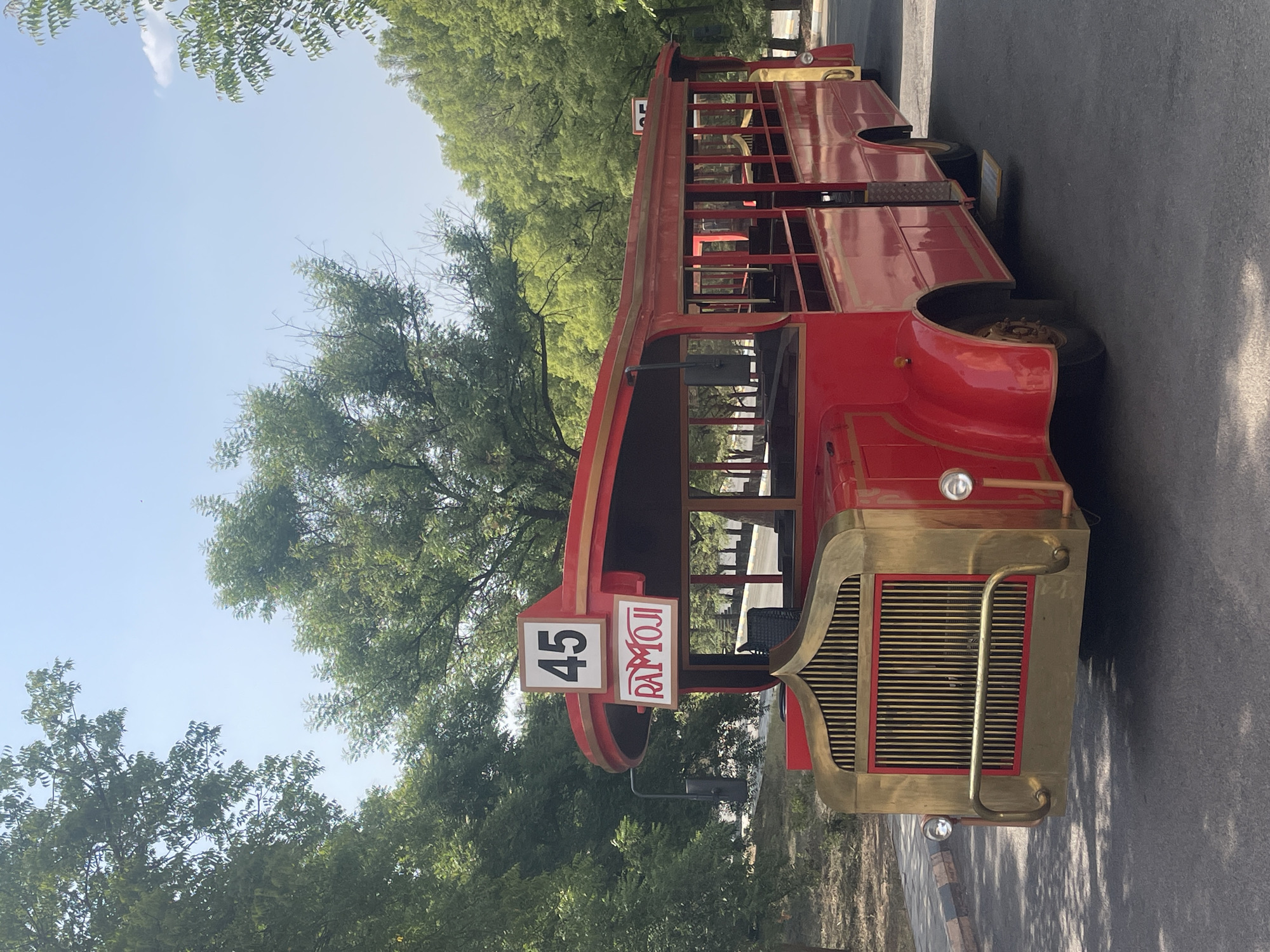 A double-decker red bus with gold-colored detailing is parked in an outdoor location. Signage on the bus includes the number "45" and the word "RAMOJI." The bus is surrounded by trees and greenery. The location is most likely Ramoji Film City in Anajpur, India. The bus appears to be a stationary prop or display rather than in active use for transportation. The surrounding area is a paved road or walkway.
