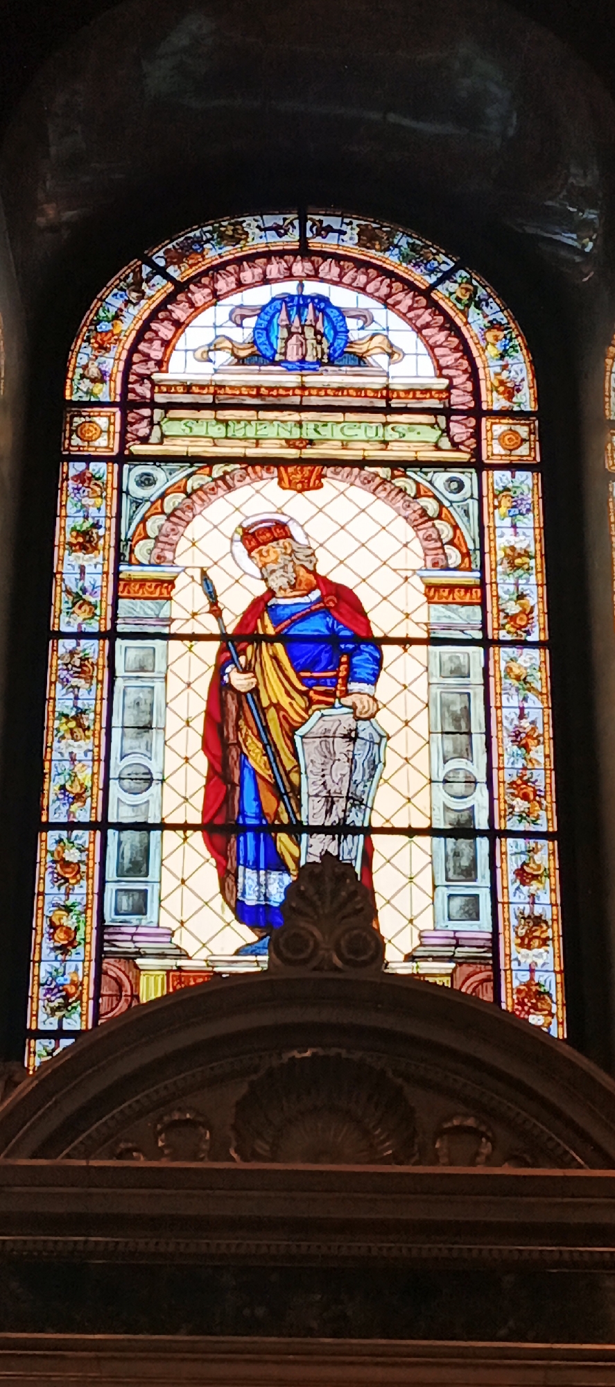 The image depicts a stained glass window featuring a figure identified as St. Henricus. The window is arched, with a decorative border of floral and geometric patterns. Above the central figure is the inscription "ST. HENRICUS." The figure is depicted with a red cloak, blue robe, and holding a spear and a shield. Behind the figure is a grid-like background with a faint representation of architectural elements. The top of a decorated structure is visible at the bottom of the image. The image was taken at Matthias Church, located in Budapest, Hungary.