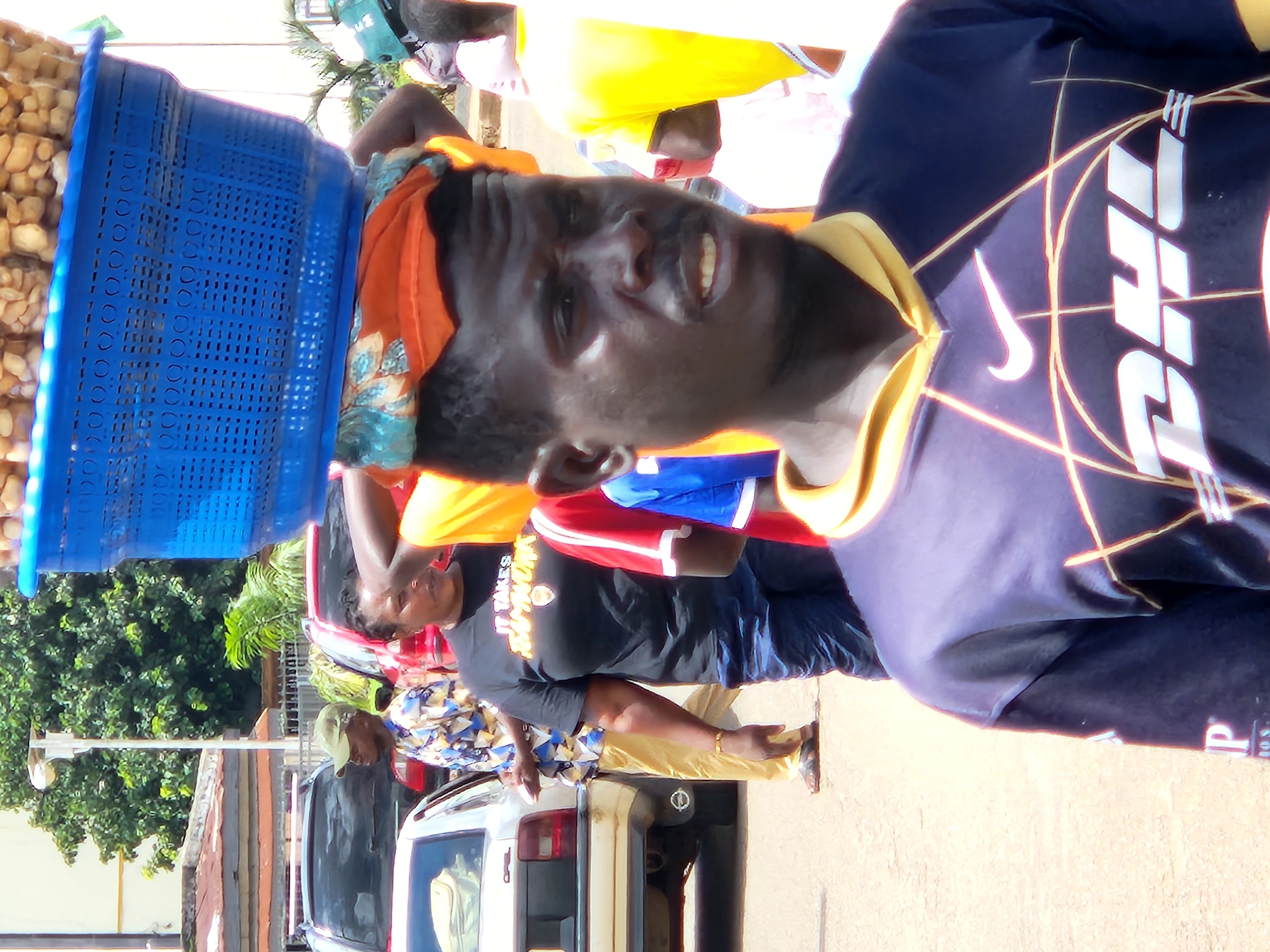 The image depicts an outdoor scene, likely a street or market, in an urban environment. In the foreground, a man with dark skin and short, dark hair is looking towards the right of the frame with an open mouth. He is wearing a dark blue t-shirt with a yellow collar and a graphic design featuring a Nike logo and the letters "DHL". He is carrying a large blue basket on his head, filled with what appear to be nuts or seeds. Behind him, a woman with dark skin and dark hair pulled back is wearing a black t-shirt with white text that reads "IT TAKES HAMMOND". She is also wearing dark blue pants. To the left of the woman, a man in a light-colored, patterned shirt and light yellow pants is walking away from the camera. He is wearing a green baseball cap. Further in the background, a white car is parked, and other vehicles and buildings are visible. The ground is paved with a light-colored surface. Trees and vegetation are present in the background, suggesting a tropical or warm climate. The lighting indicates it is daytime, with bright sunlight casting shadows.