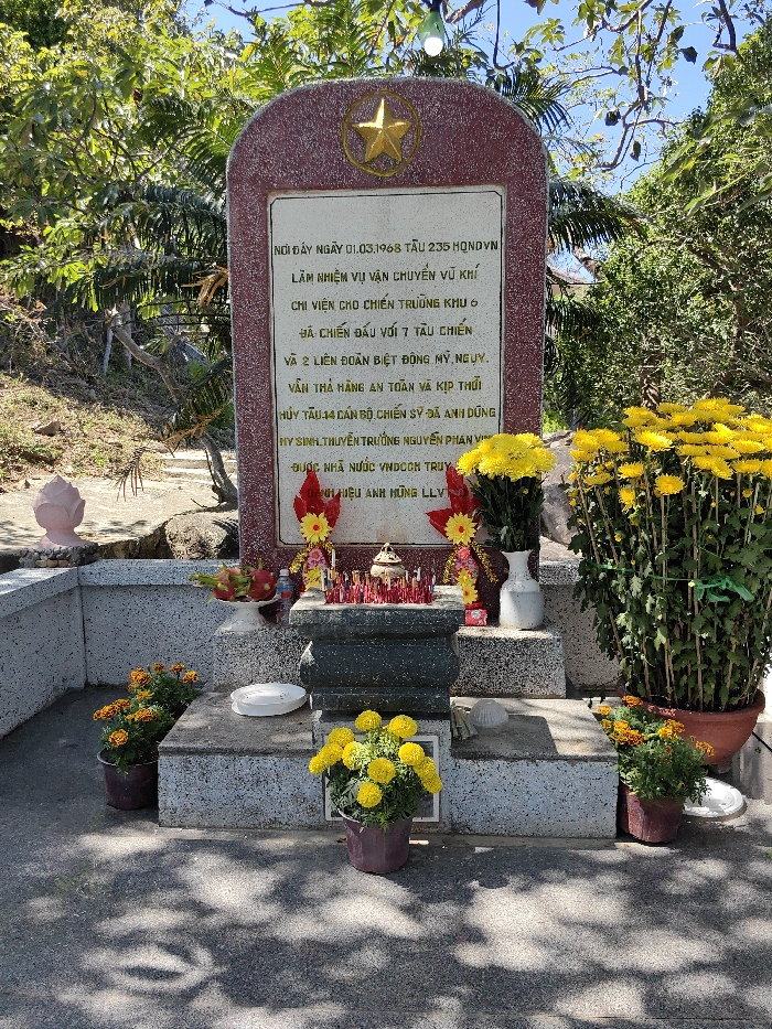 The image shows a memorial with a red stone stele marked with a gold star. The plaque commemorates the events of March 1, 1968, where vessel 235 of HONDVN carried out a weapons transport mission to support battlefront area 6. It battled 7 warships and 2 American-backed allied special forces units, managed to safely deliver the cargo, and scuttled the ship. Fourteen officers and soldiers died heroically, including Captain Nguyen Phan Vi. The memorial also states that the ship was posthumously awarded the title of Heroic Unit by the state. Flowers and offerings adorn the site.
