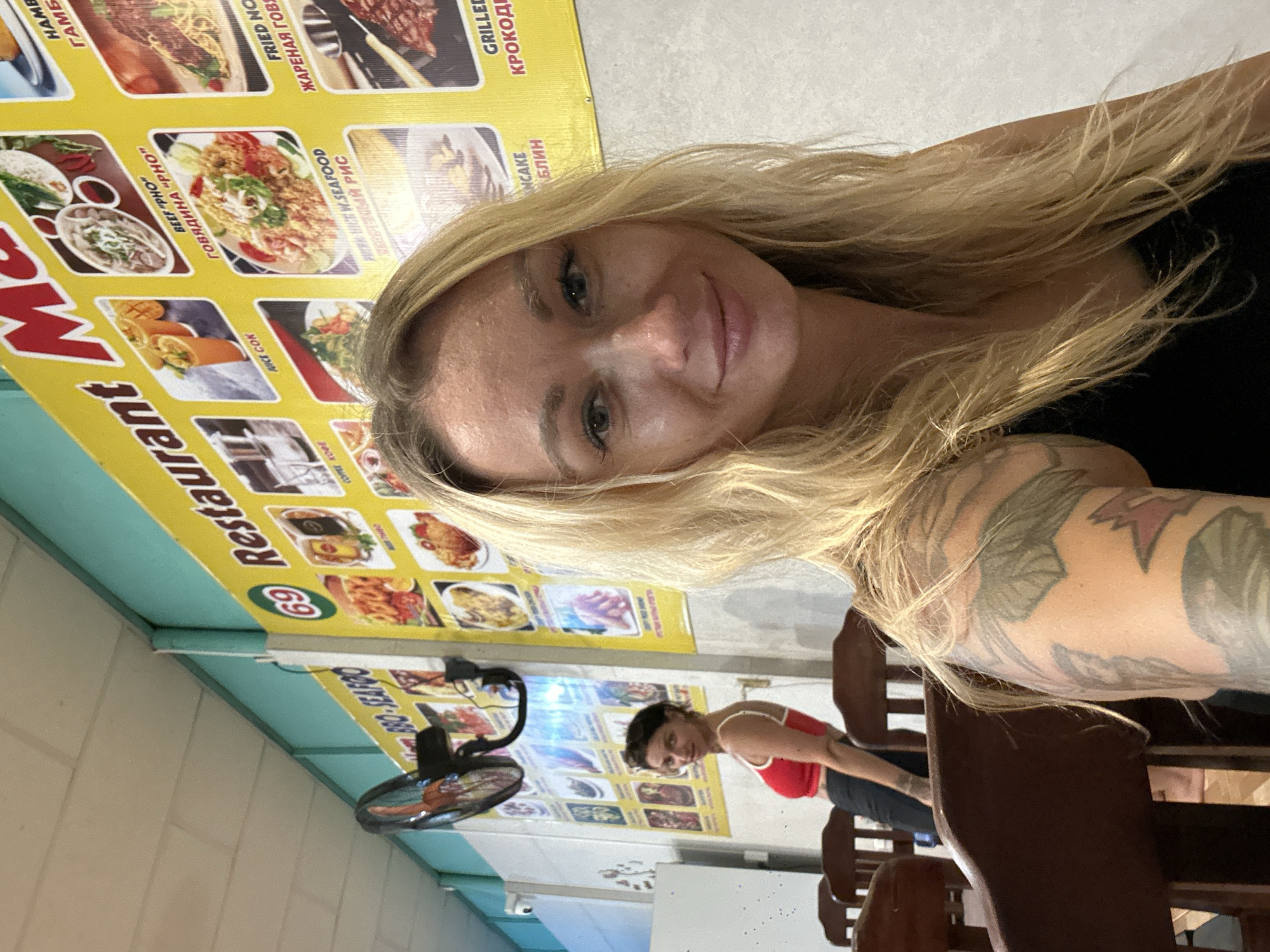 In this vertically oriented image, a blonde woman takes a selfie, her face and blonde hair dominating the foreground. She has blue eyes and a slight smile. Her arm, adorned with a large tattoo, is visible. Behind her, a menu with pictures of food is displayed on a yellow wall. The menu features items such as "BEEF PHO," "FRIED NO," and "GRILLED KROKOD." A portion of the word "Restaurant" is visible on the menu. In the background, a second woman with dark hair, wearing a red top, is seated, looking to the side. A fan is positioned on a table to the left. The setting appears to be a casual restaurant in Phan Thiết, Vietnam, indicated by the visible text on the menu which includes Cyrillic script and food items commonly found in Vietnamese cuisine. The lighting suggests it might be daytime or indoors with bright artificial lights. The overall mood seems relaxed and casual.
