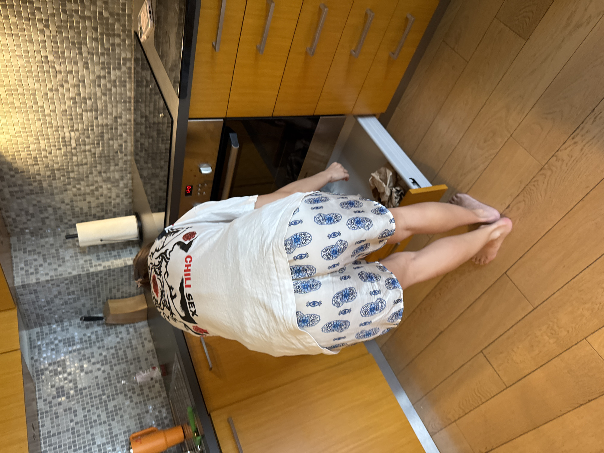 A person is in a kitchen. The person is wearing a white t-shirt with "CHILL SEX" printed on the side and white shorts with a blue pattern. The kitchen has light brown cabinets, gray backsplash, a black oven, and wood flooring. The location is in an unknown city and unknown.
