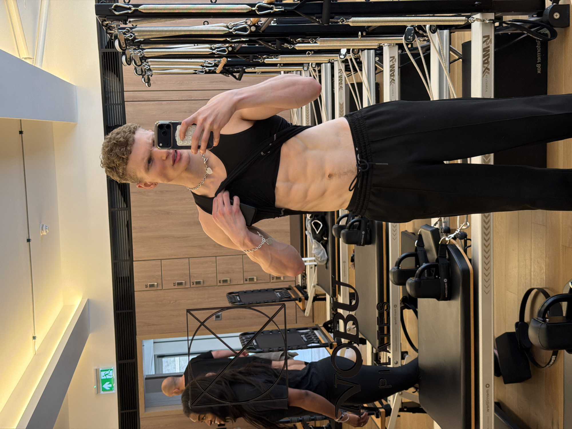 A muscular, light-skinned man with short blonde curly hair takes a selfie in the mirror of a Pilates studio. He's wearing a black tank top pulled up to expose his defined abdominal muscles, black pants, and a silver chain necklace and bracelet. In the mirror's reflection, another individual is visible, clad in black attire.
The background reveals Pilates equipment, with the "NOBU PILATES" logo visible on the machines. The Pilates studio has light wood paneling. In the reflection, an exit sign is visible, indicating the location in London, United Kingdom.