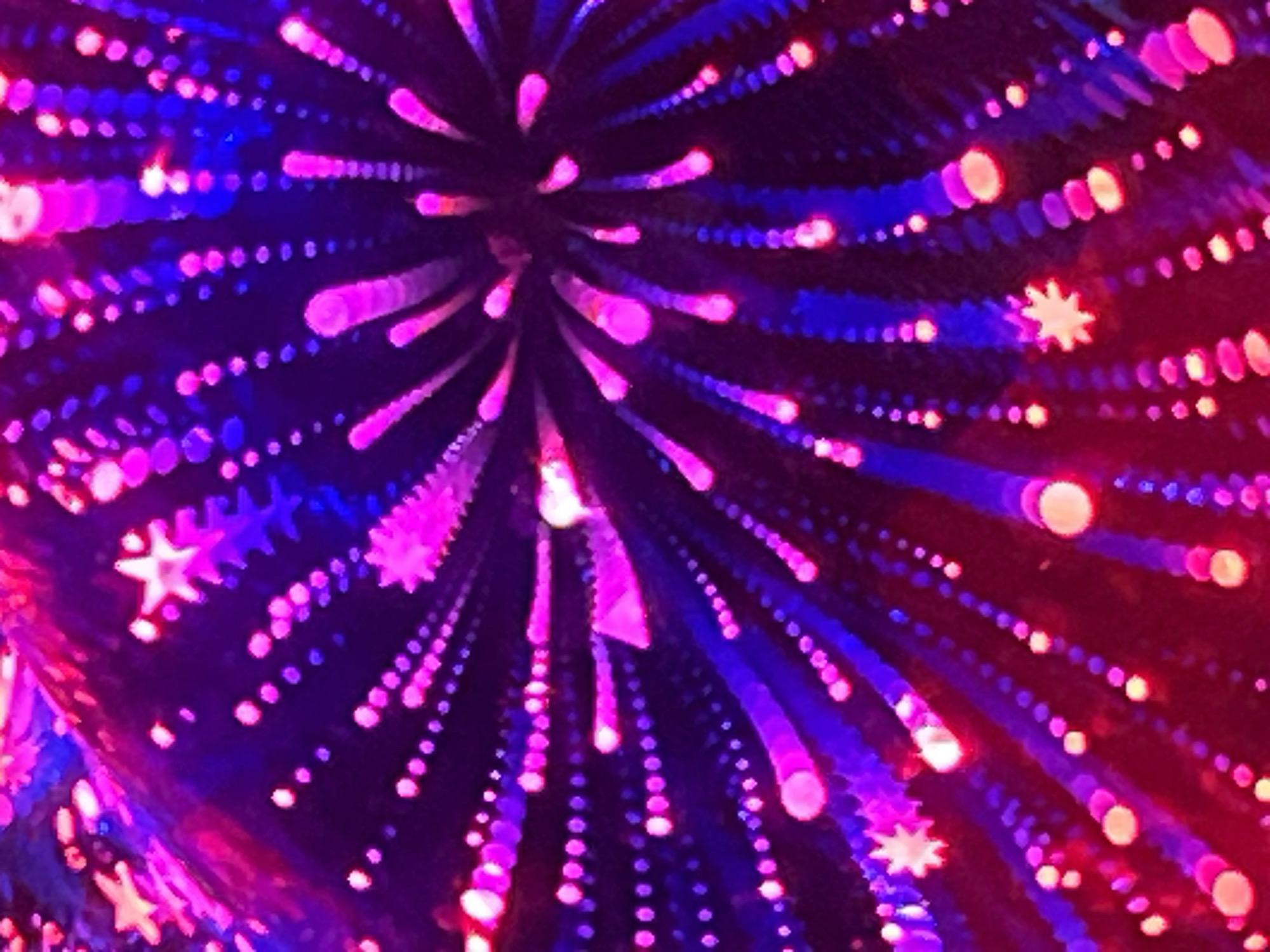 The image shows a bokeh effect with out-of-focus lights. The lights are predominantly in shades of pink, purple, and blue, creating a vibrant and energetic display. Some of the lights are shaped like stars, adding a festive element. The pattern suggests a burst or explosion of light, possibly from fireworks or a light show. The background is dark, allowing the bright lights to stand out prominently. There are no identifiable people, objects, actions, or interactions visible in the scene, and the location is unknown.