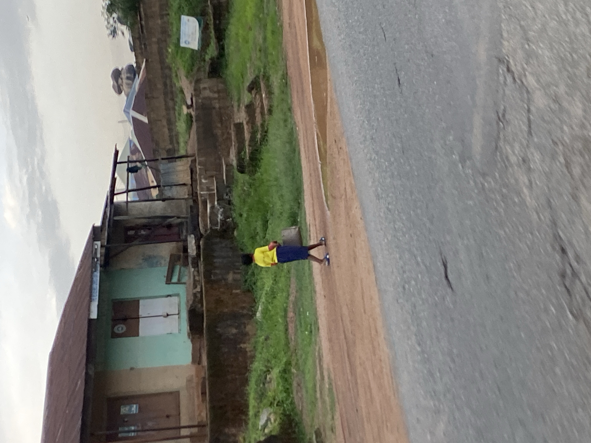 A young person wearing a yellow t-shirt and a blue skirt walks along a dirt road in front of a building in Gwong, Nigeria. The building has a rusted metal roof and is painted in sections of beige and light green. The ground in front of the building is uneven and covered with grass and dirt, with some stone steps leading up to it. A puddle of water is visible on the dirt road to the right of the person. In the background, there are other buildings, including some with red metal roofs, and a distinctive rock formation on a hill. The sky is overcast.