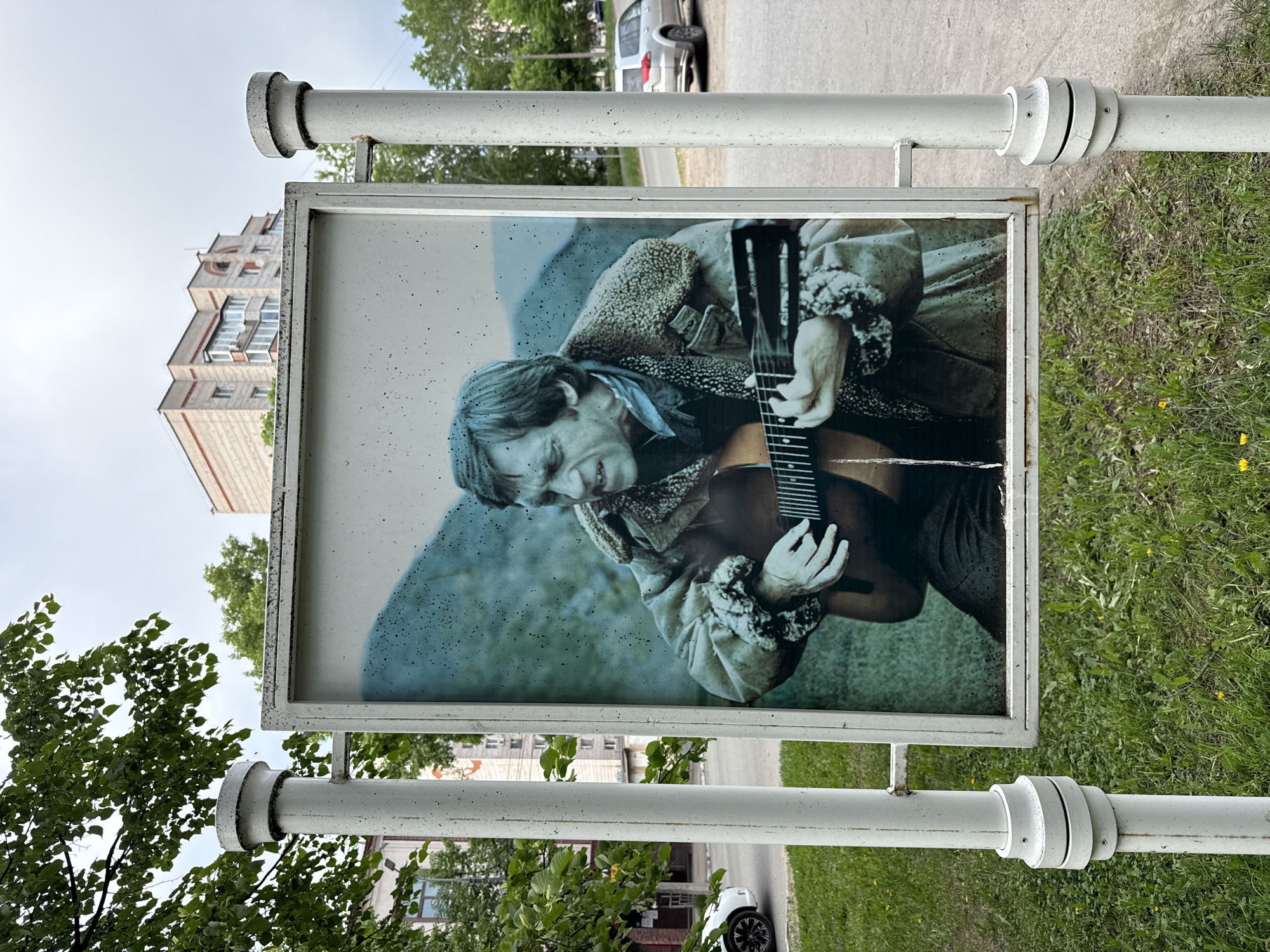 The image shows a poster of a man playing a guitar, mounted on a metal frame supported by two white metal poles. The poster's background depicts a blurred, mountainous landscape. The man in the poster has long hair and is wearing a jacket with fur lining. The poster appears weathered. The location is the city of Votkinsk, Russia. In the background are buildings, trees, a road, and parked cars. The immediate surroundings of the metal frame are grassy. The day appears overcast.