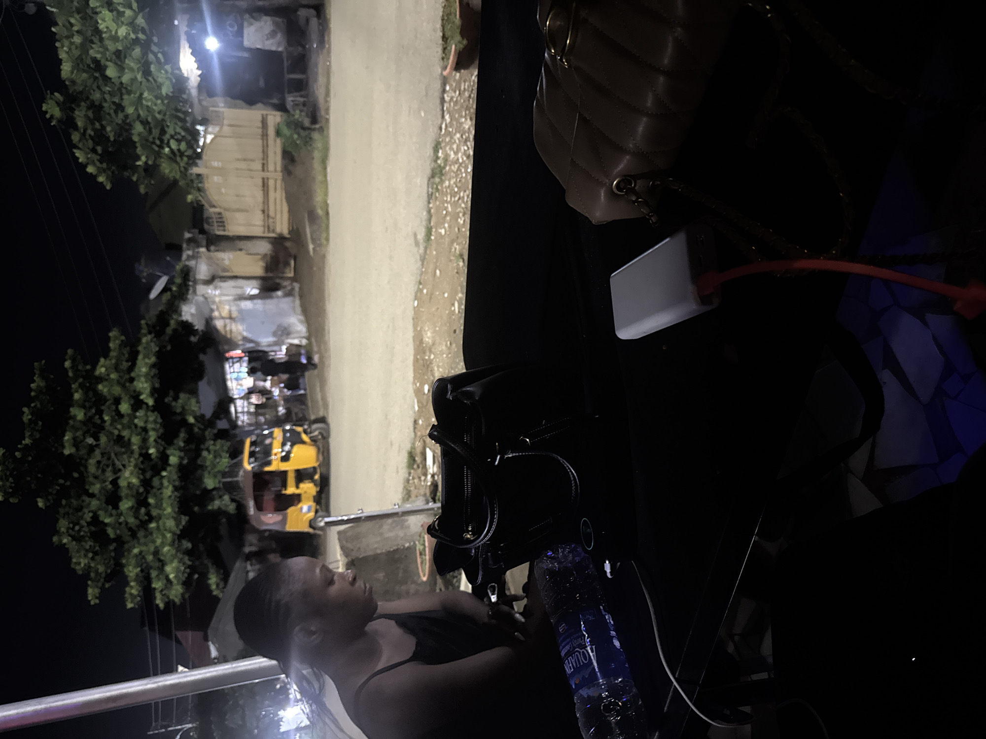 The image depicts a dimly lit outdoor scene at night, likely in Abuja, Nigeria. In the foreground, a woman with her hair in braids is seen from the chest up, looking down and to her left, with her left arm resting on her lap. She is wearing a dark-colored sleeveless top. In front of her, on a dark surface, are several objects: a partially visible black handbag, a plastic bottle of water with blue liquid, and a white power bank with a red charging cable. To the right of these items, a quilted beige handbag with gold chain straps is also visible.
The background of the image reveals a street scene at night. A yellow and red auto-rickshaw (a "Keke Napep") is parked on the right side of the street. Beyond the vehicle, there are buildings with illuminated windows and a large, leafy green tree. The street itself appears to be paved with some gravel and dirt. The overall impression is a street-side setting, possibly near a market or commercial area, during nighttime hours in Abuja.