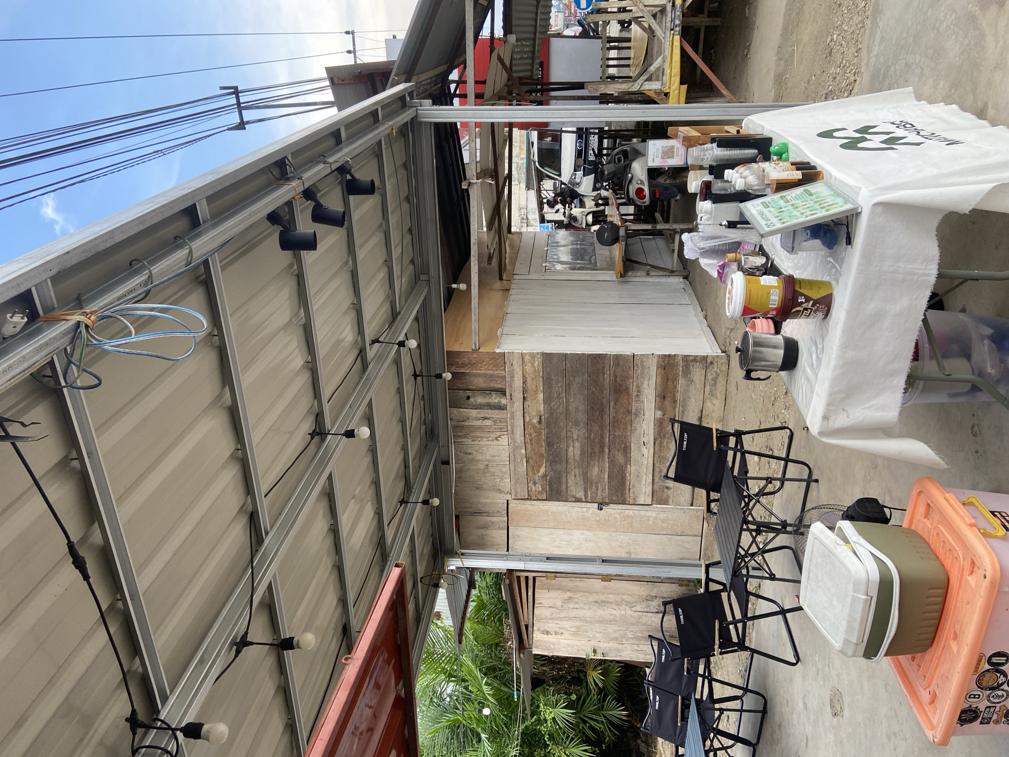 The image depicts an outdoor cafe area in Tanjung Gading, Indonesia, under a corrugated metal roof supported by a metal frame. In the foreground, a table draped with a white tablecloth displays various items, including a coffee maker, cups, bottles of liquid, and what appear to be food containers. A fan is also visible. To the left, a large orange storage bin is topped with a green and white cooler. Further back, several black camping chairs surround small tables. Behind these chairs, a rustic wooden structure forms the back wall of the cafe, with a smaller white structure to its right. In the background, several motorcycles and a white car are parked. Electrical wires are strung across the top of the frame, supported by a utility pole. Lush green foliage is visible to the left of the wooden structure. The scene is set during daylight hours with a partly cloudy sky. No people are clearly visible in the frame, though implied by the presence of a cafe setting.