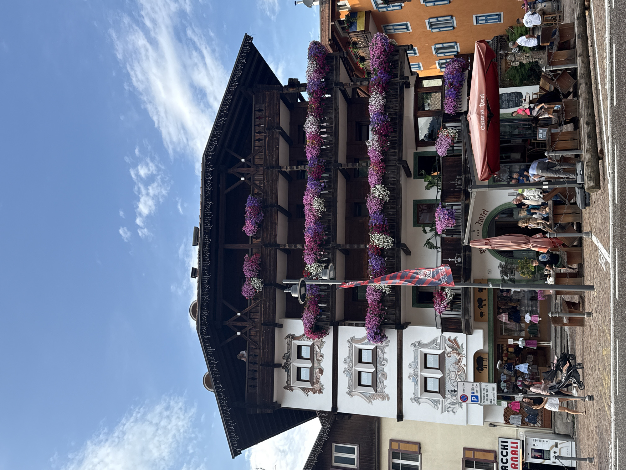The image shows a facade of a multi-story building in Moena, Italy, with traditional alpine architecture, characterized by wooden balconies and decorative painted elements. The balconies are adorned with abundant flowers in shades of pink, purple, and white. A sign indicates "Hotel" above an entrance. Below the hotel, there appears to be a shop with clothing displayed in the window and a street-level restaurant or cafe with outdoor seating. Several people are visible on the sidewalk and at the outdoor tables, some with a stroller. The building is situated on a cobblestone street. The sky above is blue with scattered white clouds. The overall scene depicts a lively street in a European town, likely during daytime.