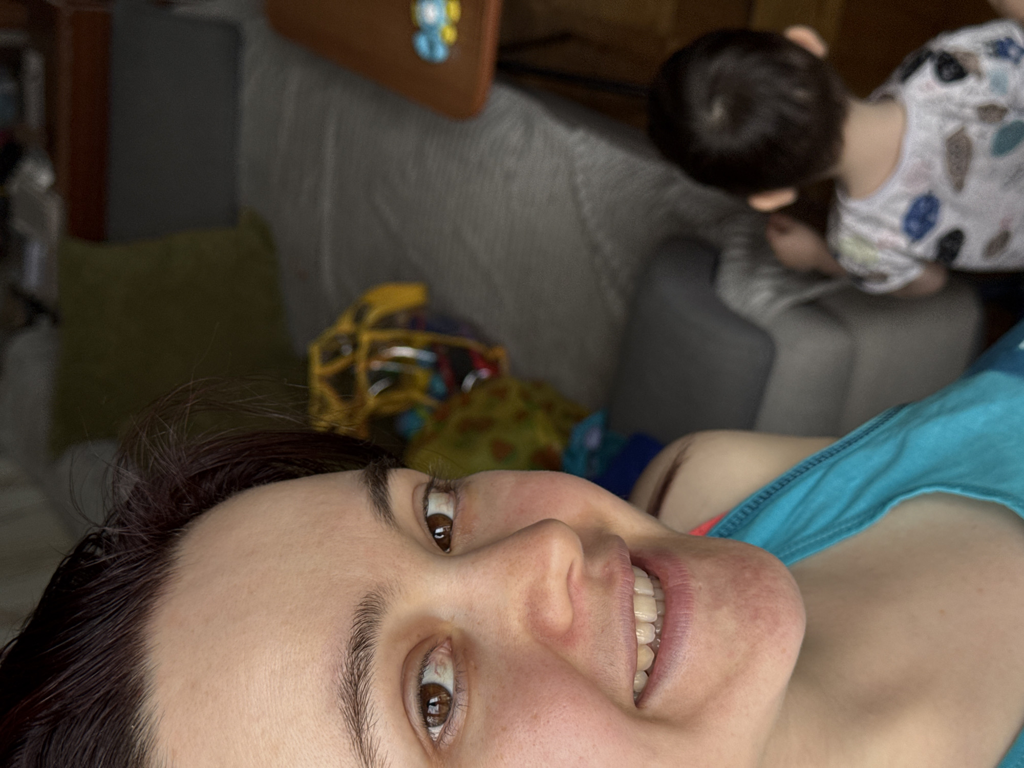 The image captures a casual indoor scene in Izhevsk, Russia, with a woman in the foreground and a child in the background. The woman is positioned in a close-up, low-angle perspective, likely lying down, with her head tilted. She has dark hair and brown eyes, and is seen smiling broadly, revealing her teeth, which suggests a cheerful or content mood. She is wearing a blue top.
In the soft-focused background, a young child with dark hair is partially visible from behind and the side, leaning over what appears to be a grey couch or sofa. The child is wearing a light-colored, patterned shirt. Other elements in the domestic setting include a yellowish cushion, a brown object with a small blue and yellow emblem, and parts of a yellow toy, possibly a helmet, in the mid-ground. The lighting indicates it is daytime. No text is visible in the media file.