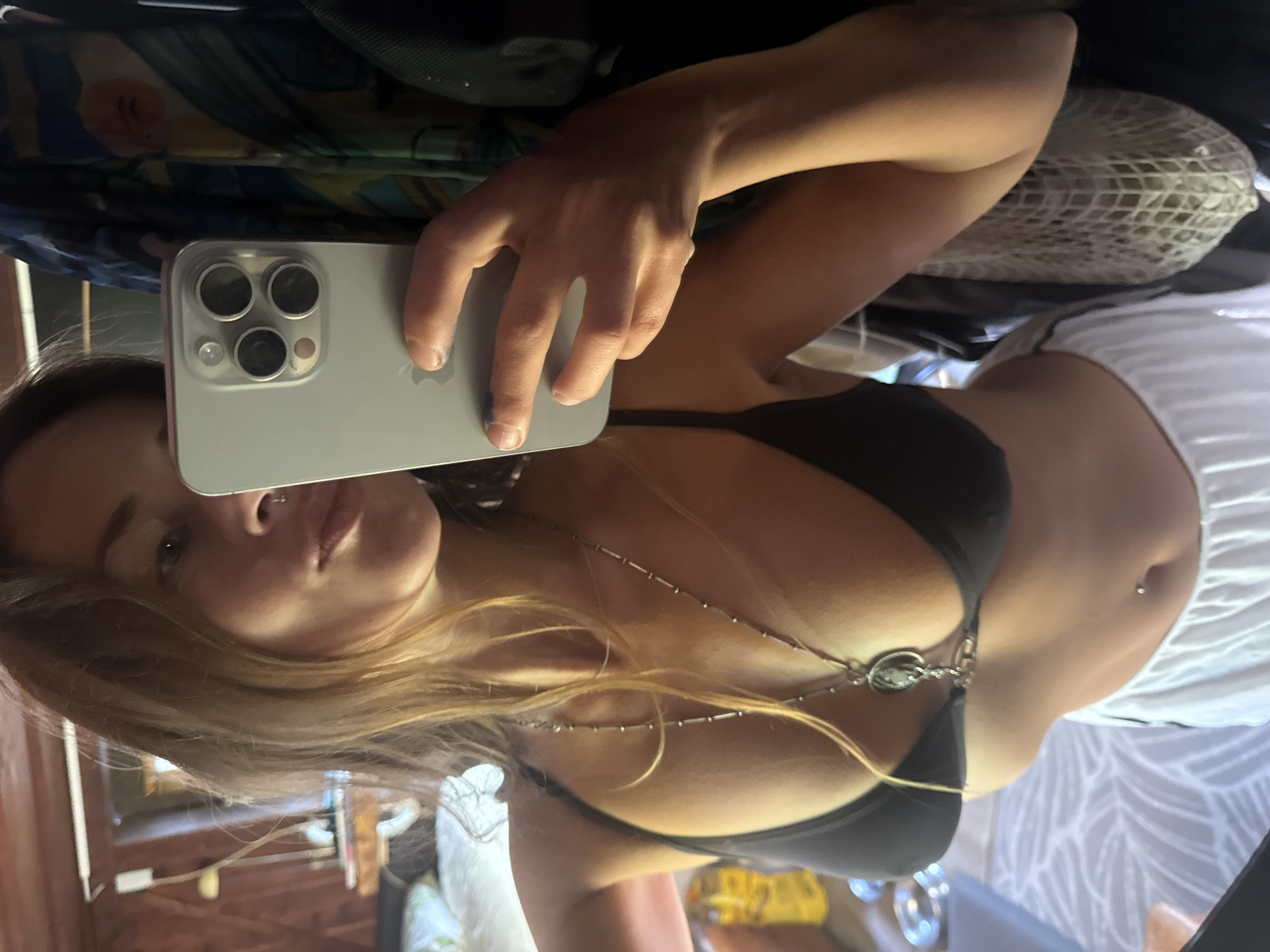 The media file is a selfie taken in what appears to be a room with wooden walls and flooring, possibly a home or a studio. The subject is a woman holding a silver smartphone with a triple-lens camera in her left hand, using it to capture her reflection in a mirror. She is wearing a black bra and white shorts, and a silver necklace with a circular pendant is visible around her neck. Her hair is long and blonde, and she has a piercing on her navel. The image is taken from a high angle, with the woman's face and upper body prominently featured. The lighting suggests it is daytime, but no specific time of day or weather conditions are discernible. The overall mood is casual and intimate, as indicated by the selfie format and the woman's relaxed posture. No visible text is present in the image.