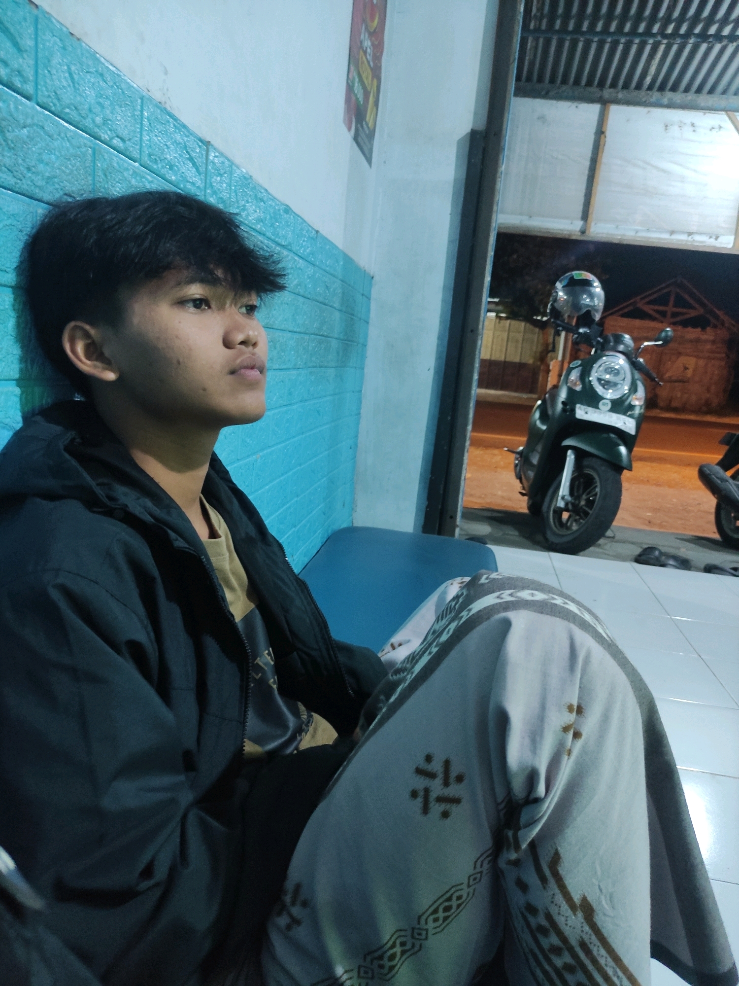 A young Indonesian male sits leaning against a textured turquoise wall in what appears to be an outdoor waiting area or covered walkway in Ngrendeng, Indonesia. He is wearing a dark hooded jacket over a tan shirt and patterned light-colored pants that are draped over his lap and legs. His hair is dark and styled forward. To his right, partially visible through an open doorway, is a dark green scooter parked on a paved surface. The scooter has a helmet resting on its handlebars. The background beyond the doorway shows a dark, indistinct area with some structures and a street with lights. The ground in the foreground is tiled. The lighting suggests it is either dusk or nighttime.