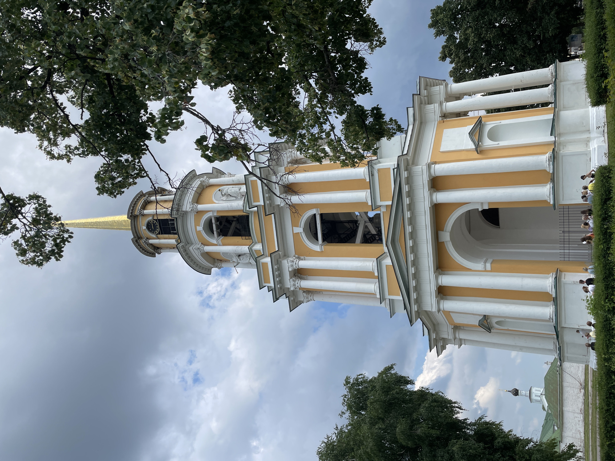 The image shows the bell tower of the Ryazan Kremlin in Ryazan, Russia. The bell tower is a tall, yellow structure with white columns and a golden spire. It is surrounded by trees and a cloudy sky. In the foreground, a group of people is gathered near the base of the tower. The scene appears to be a tourist attraction, with visitors admiring the architecture and the surroundings.