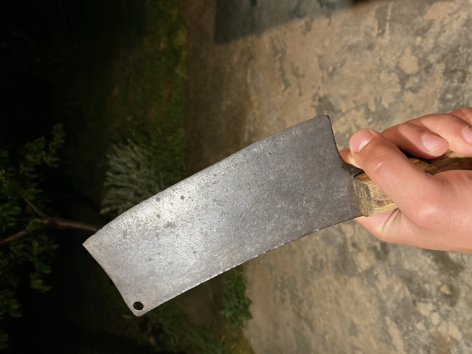 A human hand holds a weathered cleaver with a wooden handle. The blade is metal, shows signs of wear, and has a hole near its bottom corner. The background is blurred, showing foliage and a concrete surface. The scene appears to be outdoors, likely during twilight or in a dimly lit area. Konstantinovo, Bulgaria, is not discernible from the image.
