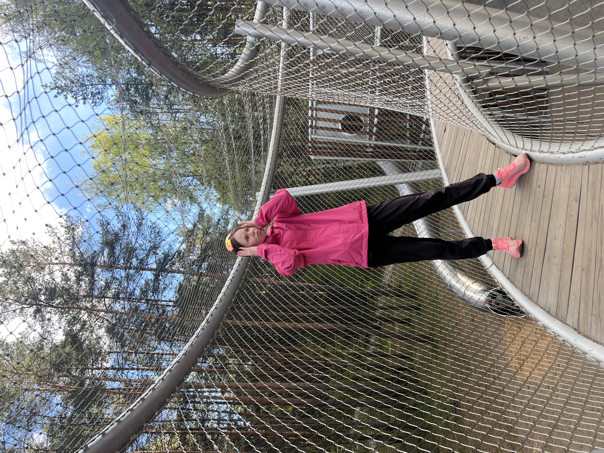 A young person stands on a wooden walkway enclosed by a metal mesh structure, possibly a canopy walk or elevated observation platform. The person is wearing black pants, a bright pink jacket, and matching pink shoes, and a yellow headband. They are holding their hands to their head, seemingly looking upwards. The background includes trees with varying shades of green foliage and a patch of blue sky. The location appears to be a forested area within or near the city of Perm, Russia.