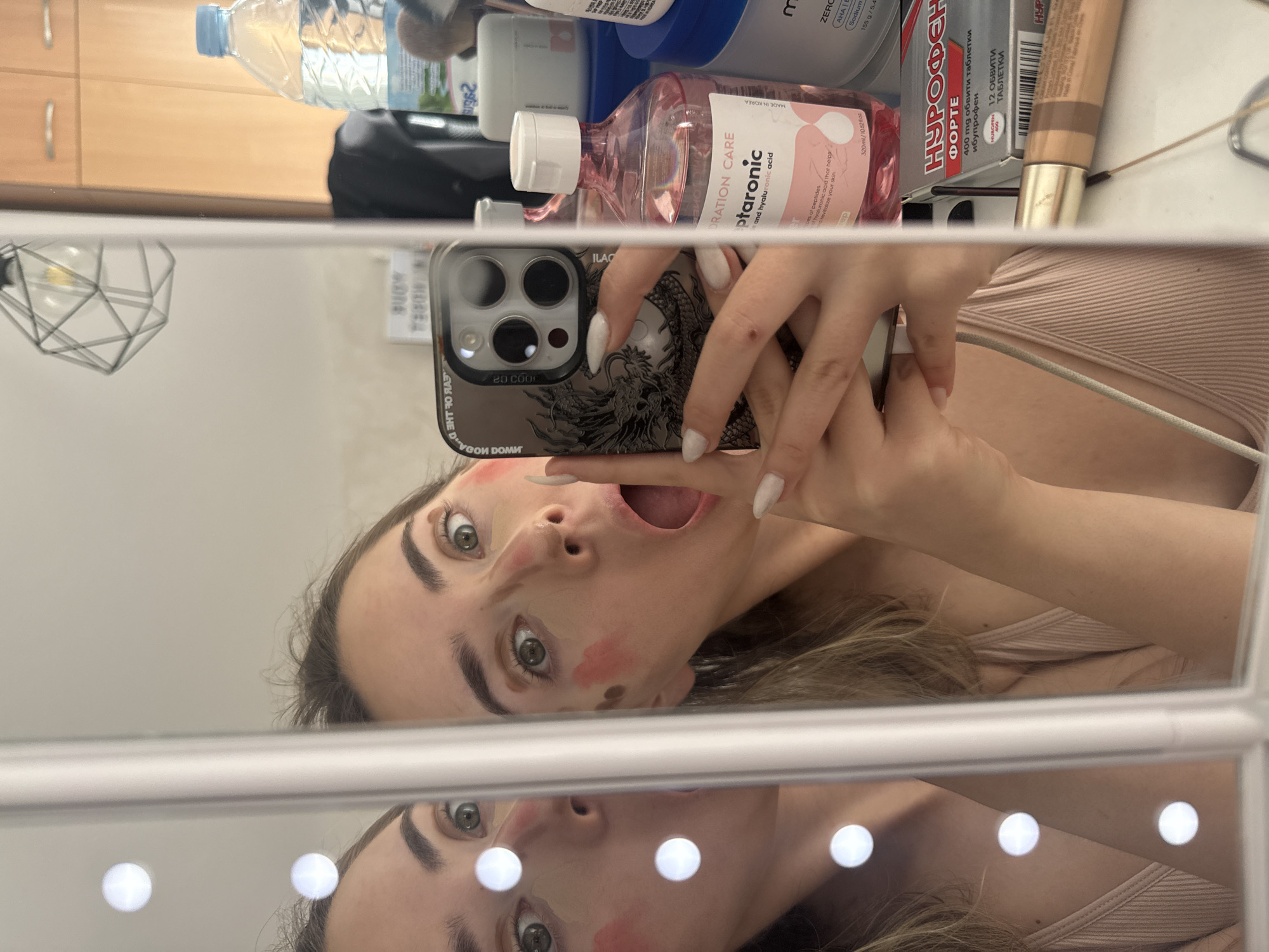 A selfie in a mirror shows a woman with light brown hair, light skin tone, and green eyes. She has cosmetic products applied to her face, specifically patches of red blush on her cheek and under her eyes. Her mouth is open, and she is holding a smartphone in front of her face with her right hand. The smartphone has a black case featuring a dragon design. Her fingernails are long and painted white.
The background reveals a bathroom setting, including various cosmetic and personal care products on a shelf. Items include a bottle of water, skin care products, and medication. A blurred reflection of a light fixture and cabinet doors is visible in the mirror above.
The text on the products includes the word "НУРОФЕН" written in Cyrillic script, as well as product information in Korean and English. These details suggest the location may be in Bulgaria, but it is not possible to specifically determine the city as Blagoevgrad.