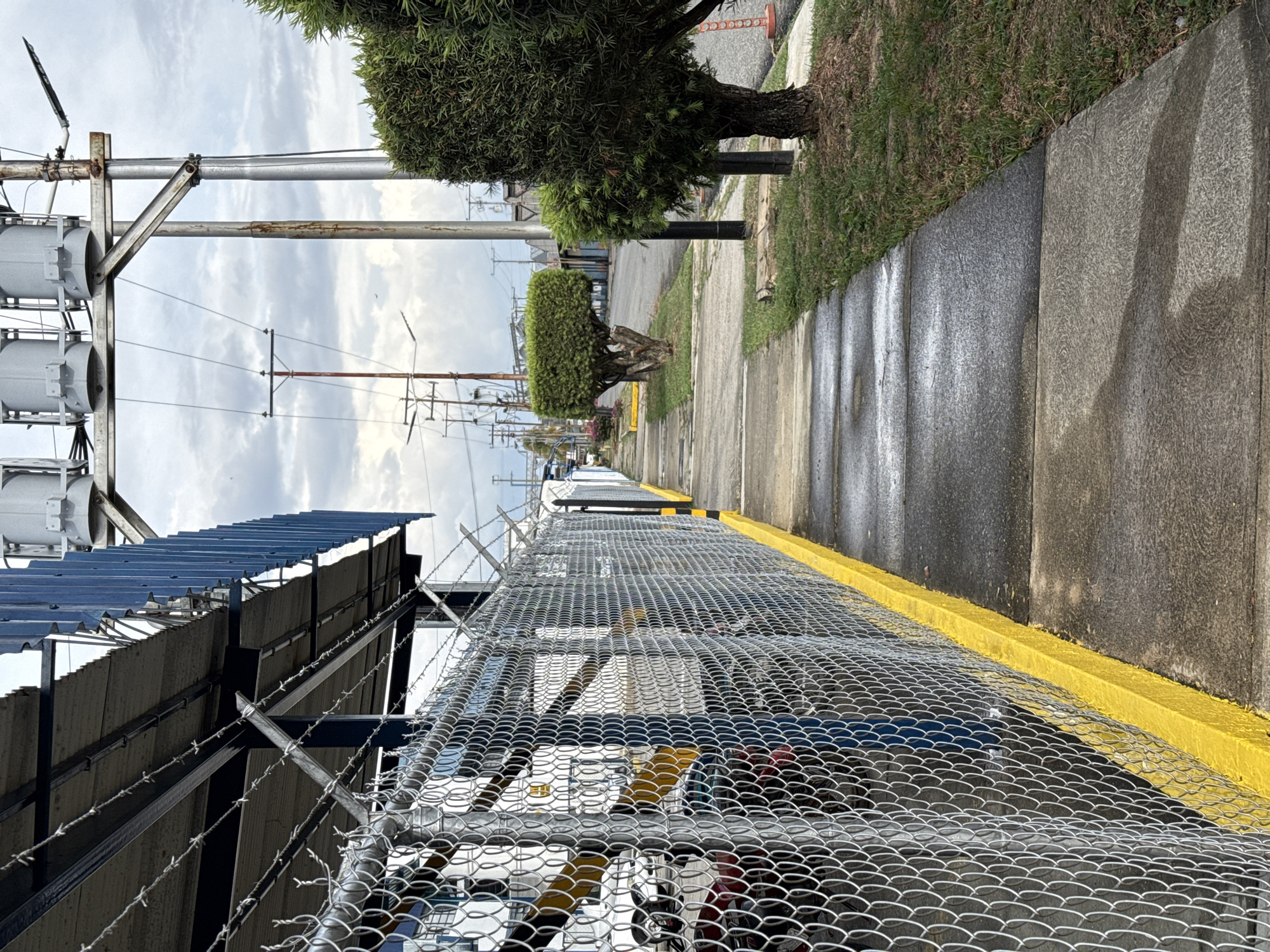 The image captures a street scene in Tinaquillo, Venezuela. A sidewalk runs along the right side of the frame, adjacent to a concrete curb painted bright yellow. A chain-link fence topped with barbed wire dominates the foreground, providing a view into an industrial area. Various vehicles, likely cars, are partially visible behind the fence. Overhead, a metal roof structure is visible, potentially belonging to a building within the fenced area.
On the left side, electrical infrastructure including poles and transformers are prominent. Further down the sidewalk, there are trees trimmed into geometric shapes. Power lines stretch into the distance. The sky overhead is cloudy. The sidewalk appears damp, suggesting recent rain. There are no people visible.