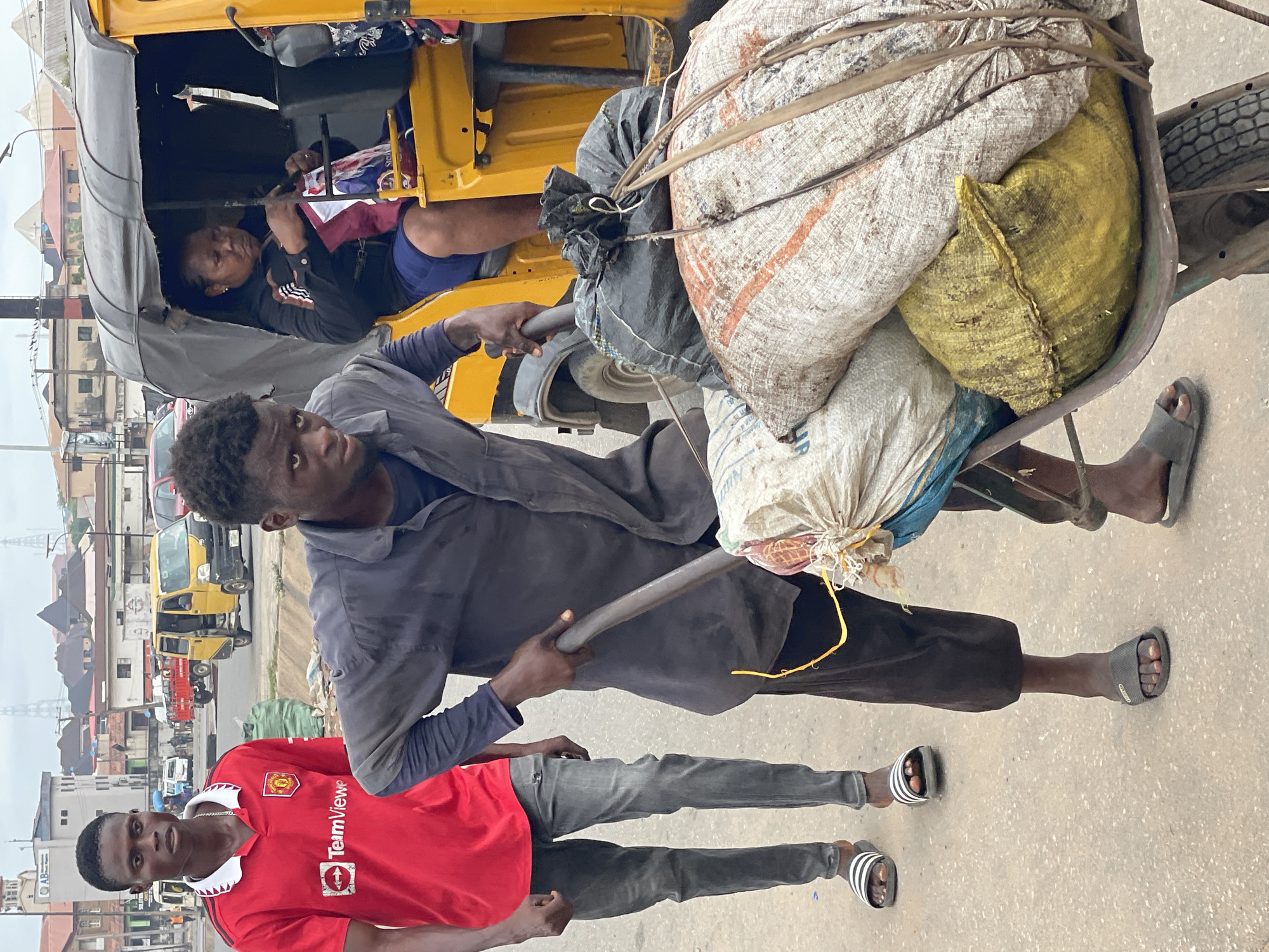 In the city of Unknown, Unknown, a man with dark skin and short hair pushes a wheelbarrow filled with sacks of goods down a paved street. He is wearing a dark, long-sleeved shirt and dark pants, and dark sandals on his feet. Behind him, another young man with dark skin and short hair stands on the left, wearing a red Manchester United jersey with a TeamViewer logo, grey jeans, and black and white striped sandals. In the background, a yellow auto-rickshaw is visible, with a woman sitting inside holding a red and white bag. Several buildings and vehicles are present in the urban environment. The sky is overcast.