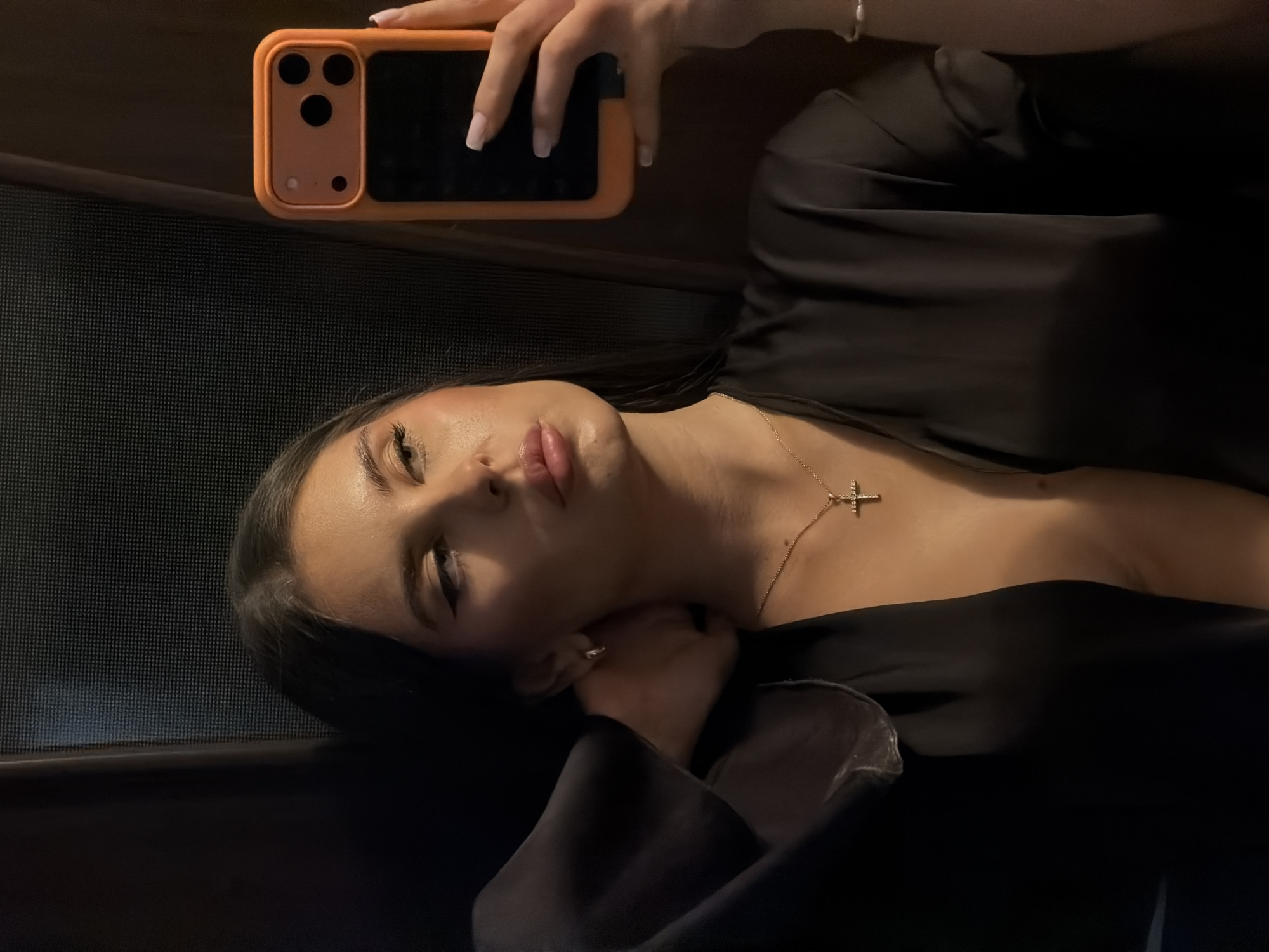 A young woman is taking a selfie. She is lying down, wearing a black top and a gold necklace with a cross pendant. She is holding an orange-cased iPhone above her head. Her fingernails are painted with white polish. The background appears to be a dark interior, possibly near a window with textured glass. There is no indication of the location being Krasnoyarsk, Russia.