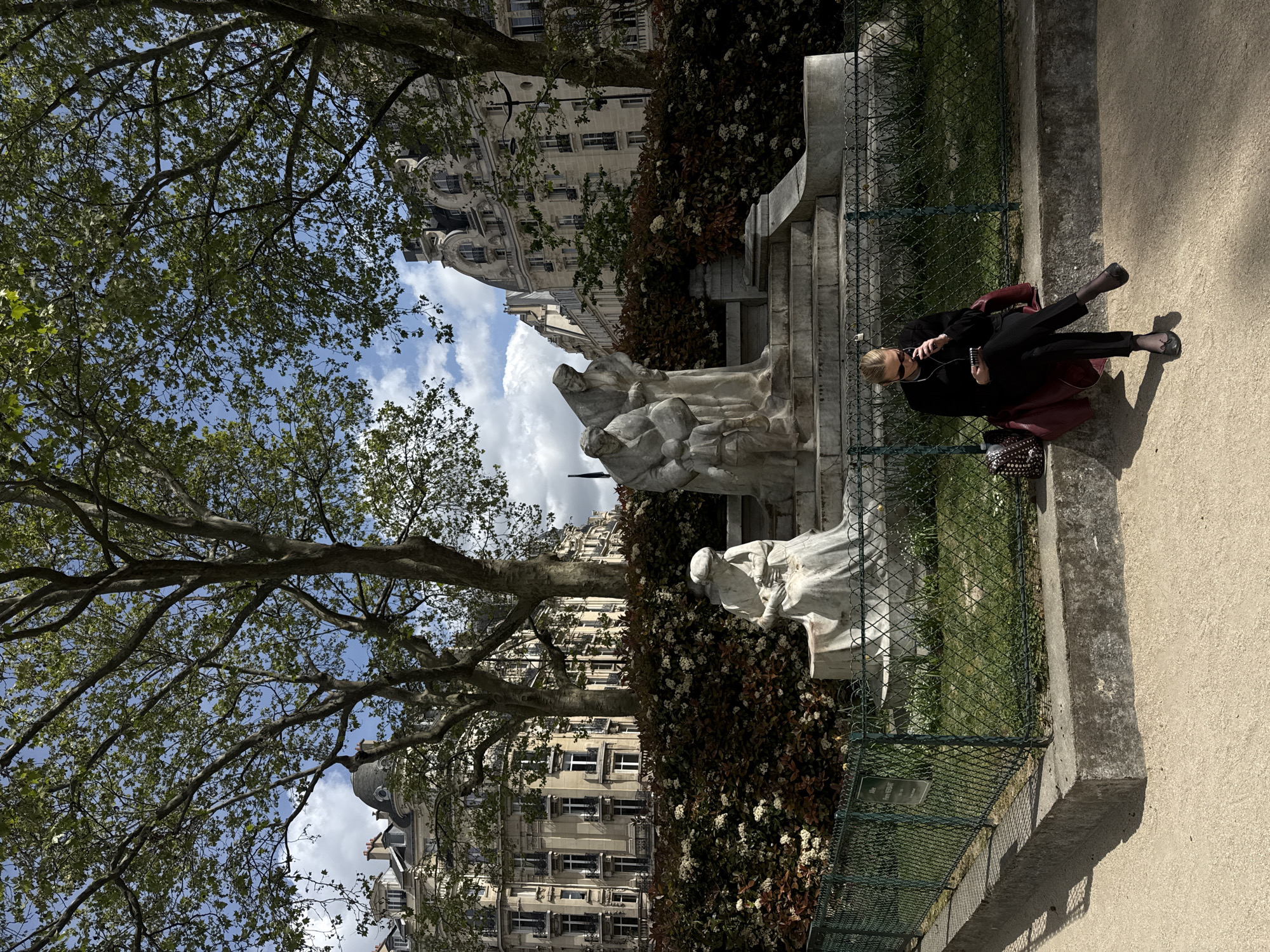 A daytime scene in a park in Paris, France, shows a woman resting on a concrete ledge next to a series of white stone statues. The image is oriented vertically but depicts a horizontal landscape.
In the mid-ground, a woman with blonde hair, wearing a black top, dark trousers, and black ballet flats, is seated on a low concrete wall or ledge. She is holding a small dark object, possibly a phone, and appears to be wearing wired earbuds. A red bag is placed on the ledge beside her, and a dark studded handbag rests on the ground. To her left, behind a green metal fence, a group of white marble or stone statues depicting several figures is visible, with another individual statue further to the left. These statues are positioned amidst a dense, dark green and reddish-brown bush with small white flowers.
The background features classic Parisian Haussmannian buildings with light-colored facades, numerous windows, and ornate details, including a dome visible on one building to the far left. Above the buildings, the sky is partly cloudy with patches of clear blue. Large trees with green foliage and bare branches frame the top and left sides of the scene, suggesting a spring or early summer day.
The foreground consists of a paved path or dusty ground, with the concrete ledge and the green metal fence running horizontally across the lower part of the image. The sunlight casts shadows from the fence and the person onto the ground, indicating bright daylight. A small sign attached to the fence near the statues is visible, with the text "ALLEE RENE HERBART". The overall impression is one of a tranquil urban park setting.