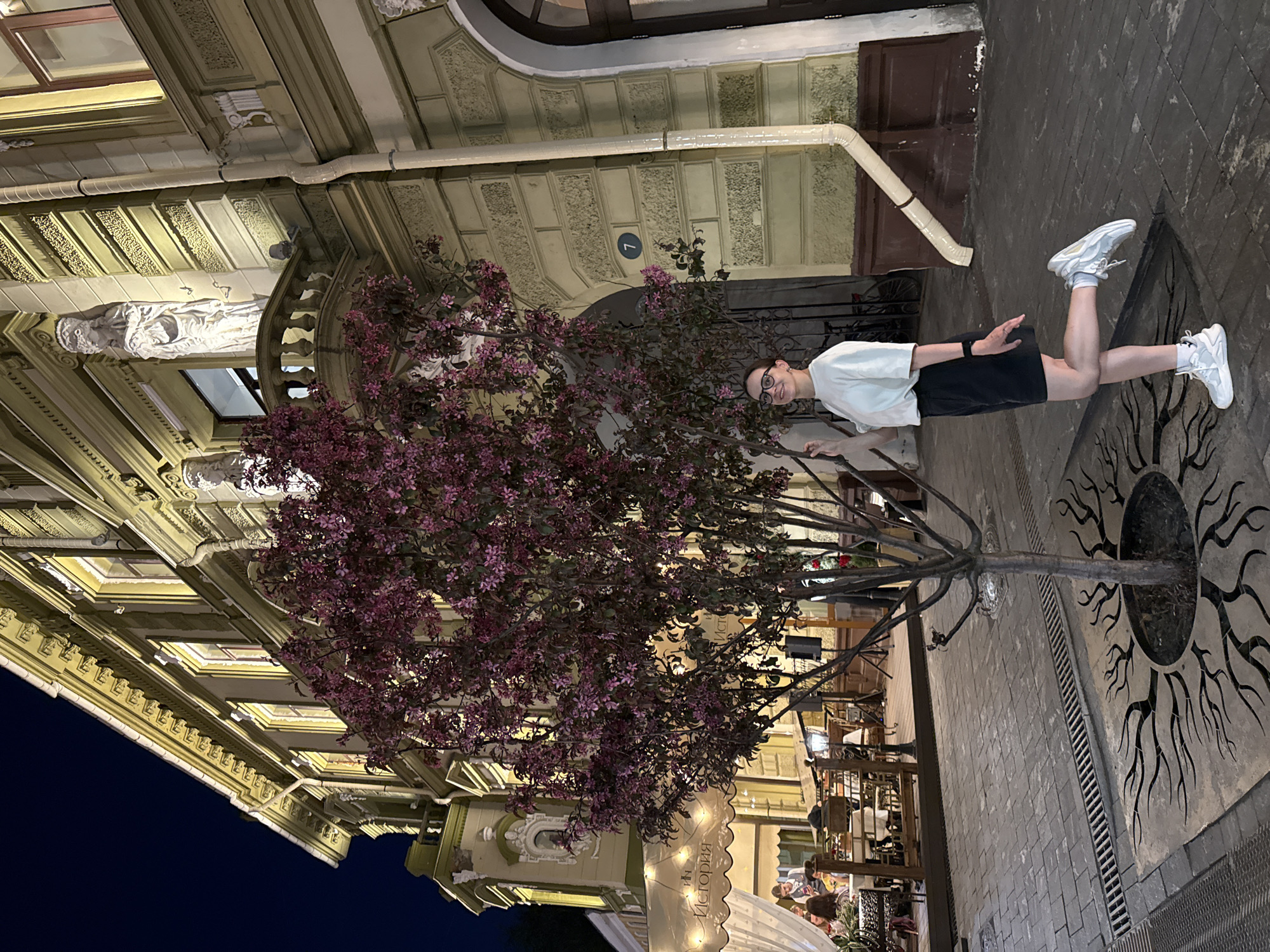 A young person in a white t-shirt, black shorts, and white sneakers is standing on the sidewalk next to a decorative metal tree sculpture in Nizhny Novgorod, Russia. The background features a tall, ornate building illuminated by lights. The sidewalk is paved with bricks. People are visible seated at tables under awnings in the background. The sky is dark. A sign with the word "История" (History) is visible in the background. A metal grate is near the tree sculpture. The building has statues and decorative moldings. A metal gutter runs along the building. A sign with the number "7" is visible on the building.