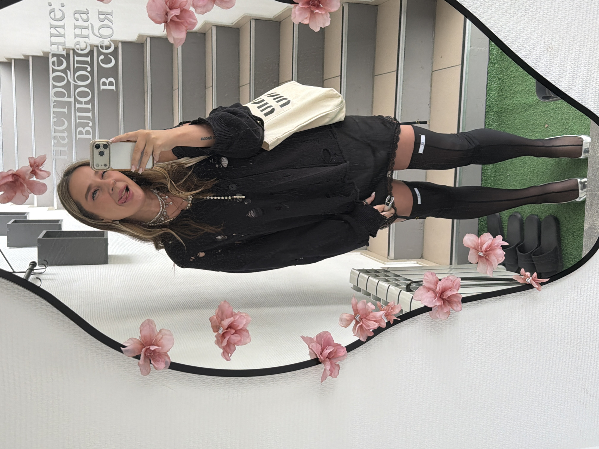 A young woman captures a mirror selfie indoors, revealing a playful demeanor. She stands in front of a large, irregularly shaped mirror adorned with artificial pink flowers. Her blonde hair is parted to the side, and she grins widely, sticking her tongue out with a slight wink. She holds an iPhone with a light-colored case up to her face, reflecting her image.
She is dressed in a casual yet edgy outfit, featuring a distressed black jacket worn over a black top and a black mini-skirt with a lace trim. Her legs are covered in sheer black thigh-high stockings with a distinct white label visible on the upper thigh, and she wears silver pointed-toe shoes. Around her neck, multiple strands of pearl necklaces are visible. She also carries a light-colored tote bag with black stylized text on it, slung over her shoulder. A tattoo is visible on her left wrist.
The mirror reflects the interior surroundings, suggesting a modern public or commercial space, possibly in Chaikovsky, Russia. In the background, a set of light-colored stairs ascends, and to the right, a section of green artificial turf is visible, featuring a pair of black sandals or slippers placed on it. Grey rectangular planters and a gridded surface are also reflected in the lower left.
Prominently displayed in large white Cyrillic letters on the mirror's surface is the text: "Настроение: влюблена в себя," which translates to "Mood: in love with myself." A smaller text, "LEA," is visible on the white label of her right stocking. The overall atmosphere conveyed is one of self-confidence and lightheartedness.