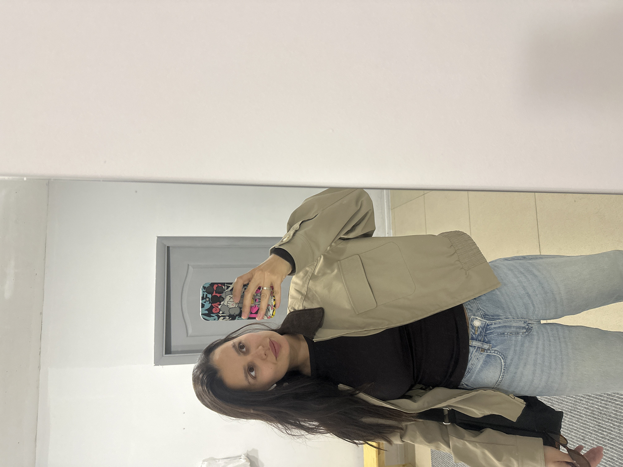 A young woman is captured in a mirror selfie, holding a smartphone to take her own picture. She has long, dark hair parted to her left and a neutral to slightly serene expression, looking directly at the camera in the reflection.
She is dressed in a casual outfit consisting of a light beige or khaki bomber-style jacket over a black long-sleeved top and light wash blue jeans. A ring is visible on her left hand, which is holding the phone. The smartphone has a distinct and colorful case featuring a pattern of abstract shapes and what appear to be KAWS-like cartoon characters in various bright colors like red, yellow, blue, and black.
The setting appears to be indoors, possibly a hallway or a fitting room, as indicated by the large mirror. In the reflection behind the woman, a light-colored wall is visible, along with a doorway framed in gray, featuring a decorative panel. The floor reflected in the mirror shows light-colored tiles and a portion of a dark and light striped rug or mat. The overall lighting is even, suggesting an indoor environment. No specific time of day or weather conditions are discernible. The provided location context is Chaikovsky, Russia.