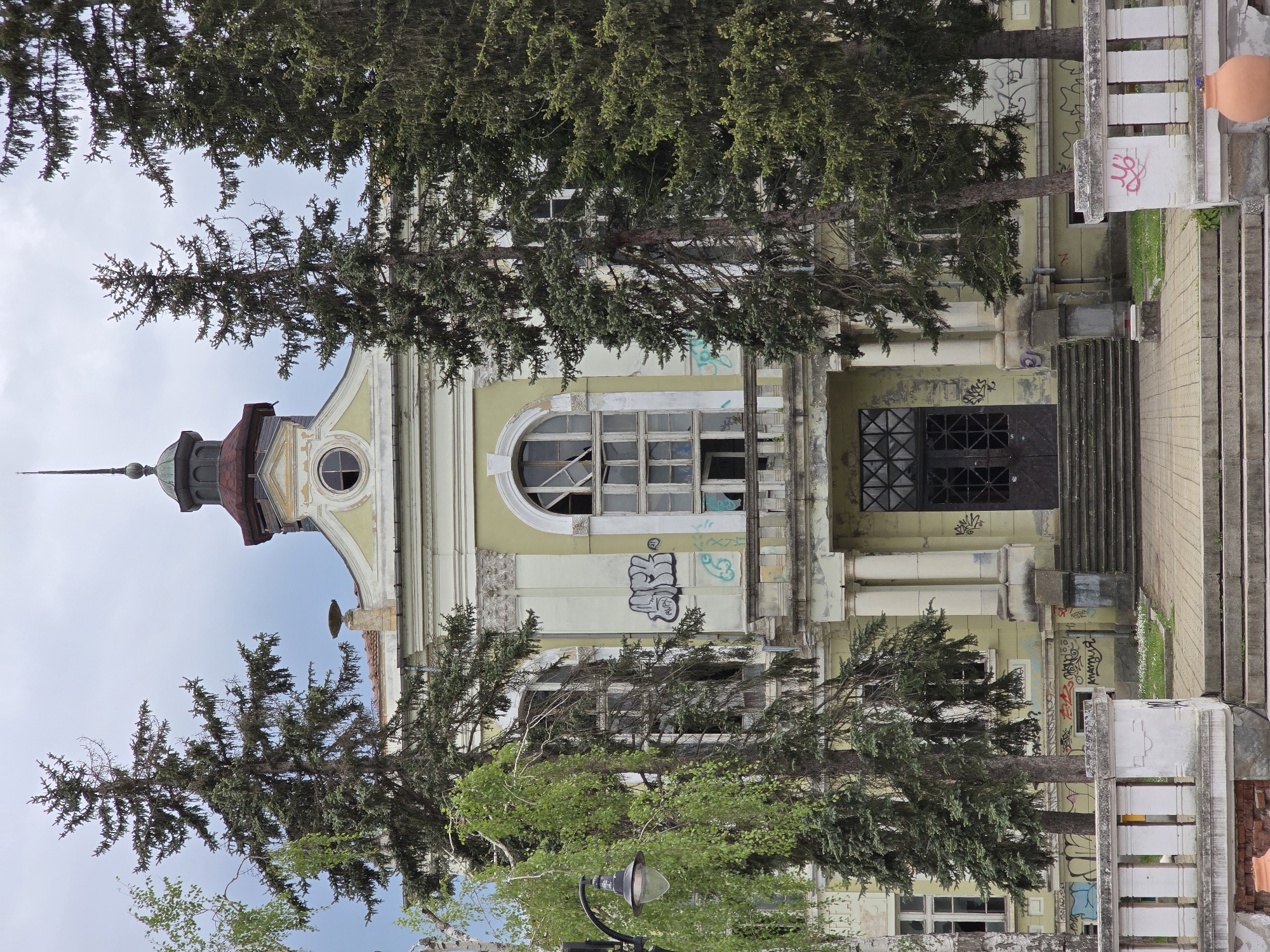 The image shows a building with a yellow facade and a tower, partly obscured by tall evergreen trees. The architecture suggests a historical building, but it appears to be in disrepair with some graffiti on the walls. The entrance is flanked by columns and leads to a set of stairs. The location is in the city of Unknown, Unknown.