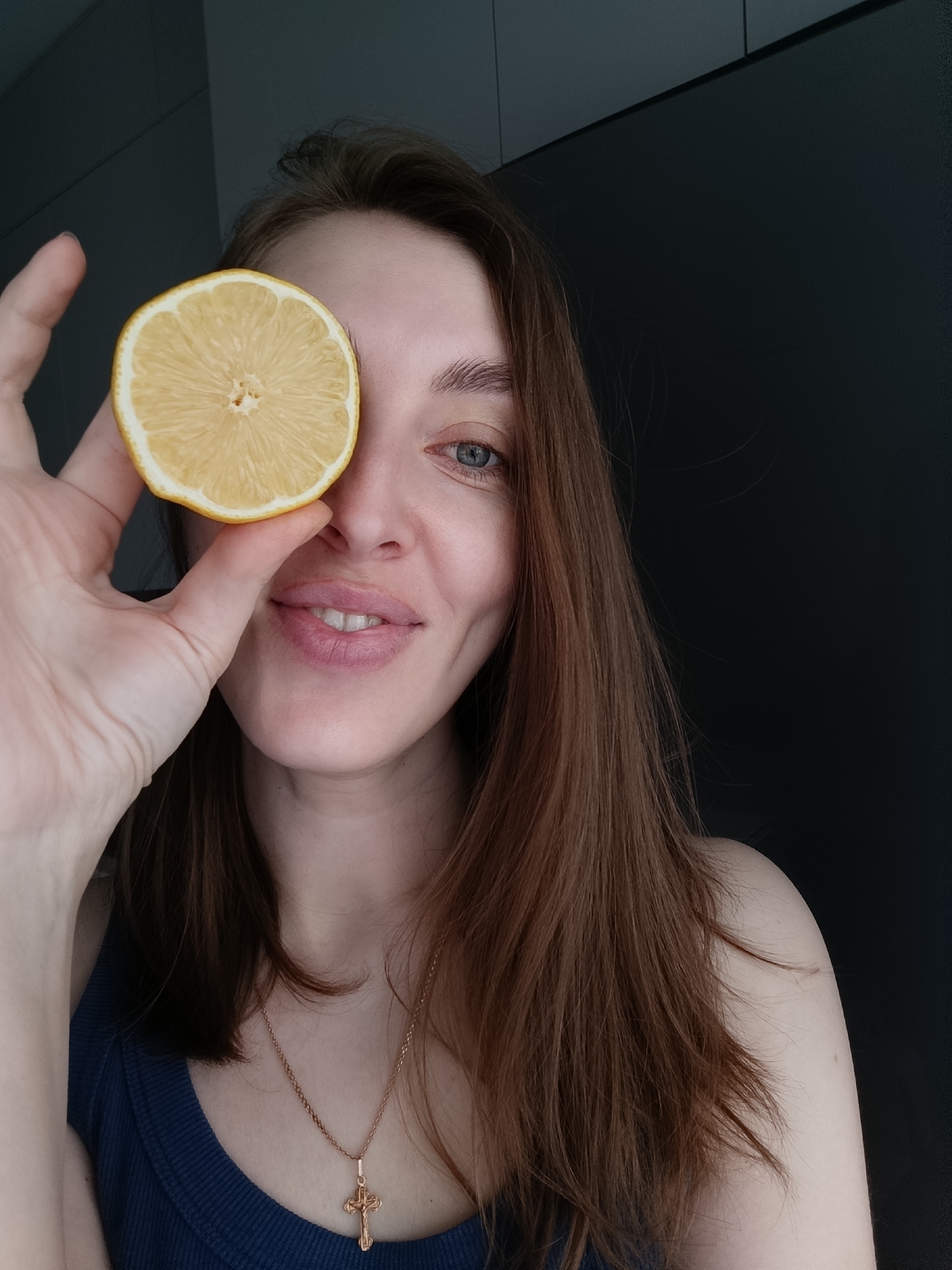 A fair-skinned woman with auburn hair holds a lemon slice over her left eye, smiling at the camera. She wears a dark blue tank top and a gold cross necklace. The background is a dark gray wall. The location is Chaikovsky, Russia.