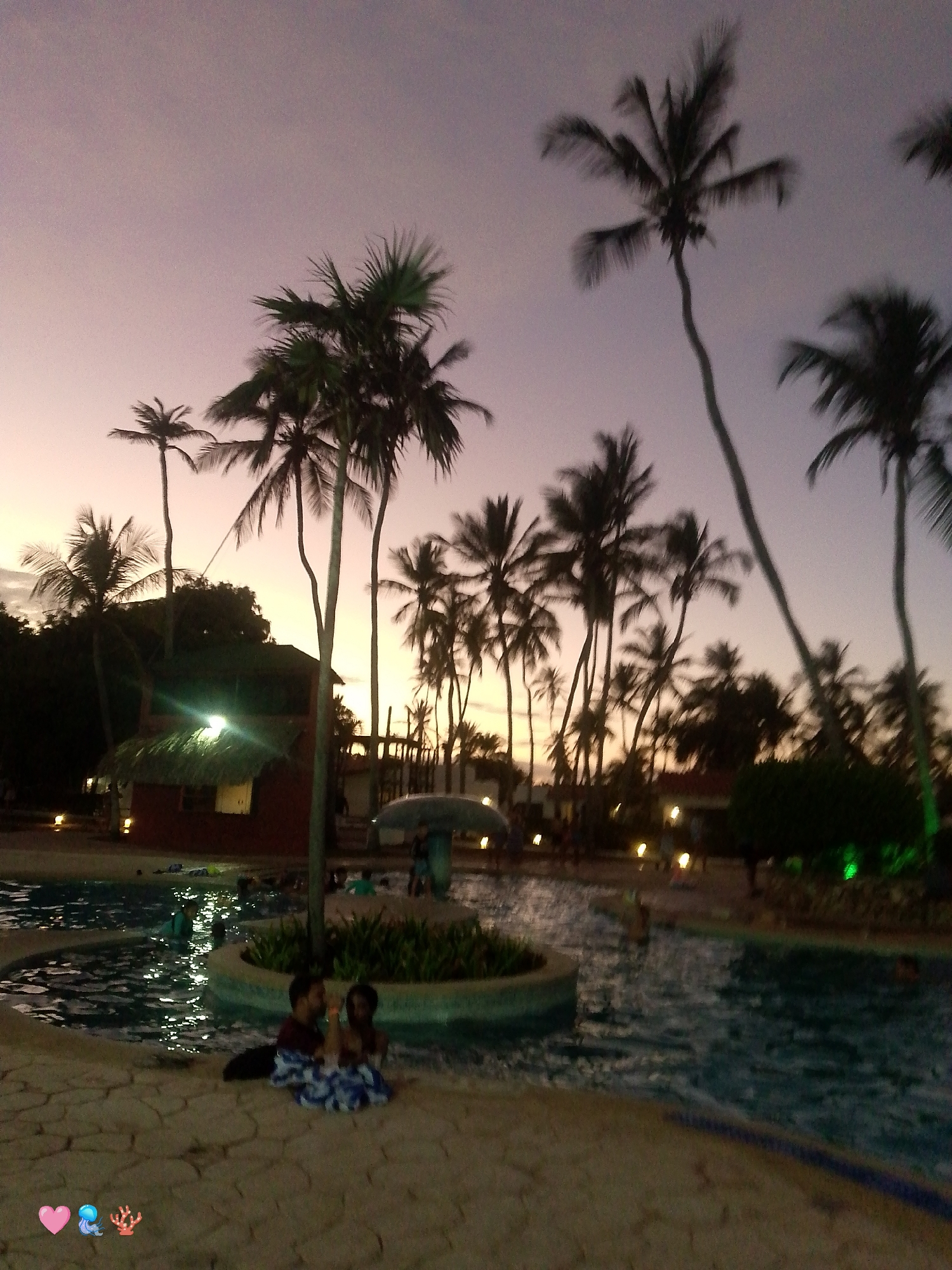 The image shows a nighttime scene at a resort in Playa El Agua, Venezuela. A swimming pool takes up a large portion of the foreground, with two children sitting on the pool deck near the edge of the water, covered with a patterned blanket. In the middle distance is a small island in the pool with plants. Behind the pool are palm trees silhouetted against a twilight sky with hues of pink and purple. A building with a thatched roof and lit windows is visible on the left side of the image. Dimly lit lamps are scattered around the pool area. Several people are in the pool and around the area.