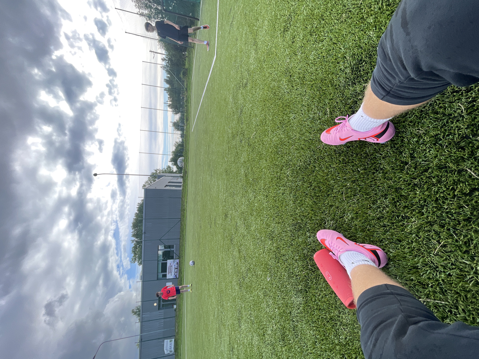 This is a first-person perspective image taken on a synthetic turf field, likely in or near Jakai, Lithuania. In the foreground, the legs and feet of the photographer are visible, wearing black shorts, white socks, and bright pink soccer cleats. To the left of the photographer's feet is a red cylindrical object, possibly a speaker or a portable charger.
Further on the field, two individuals are present. One, wearing a red t-shirt and dark shorts, stands near the center of the field. Another individual, wearing a black t-shirt and black shorts, is positioned further down the field, facing towards the right side of the frame. A soccer ball is visible on the grass between the two individuals.
In the background, a grey building with signage is located on the left side of the image, and a chain-link fence encloses the sports field. Trees and a cloudy sky are visible beyond the fence. The overall environment suggests an outdoor sports facility.