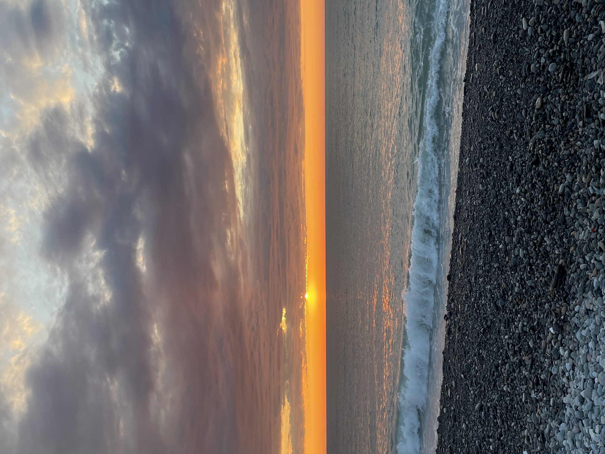 The image captures a sunset at a rocky beach in Batumi, Georgia. The sky is divided into thirds: the left side displays grey and white clouds, the center shows the sunset glow in orange, pink, and yellow, and the right side is the sea. The setting sun reflects on the water's surface. Waves are crashing on the shoreline. A pebbly beach is visible on the right edge of the frame. The image is oriented vertically. There are no people visible.