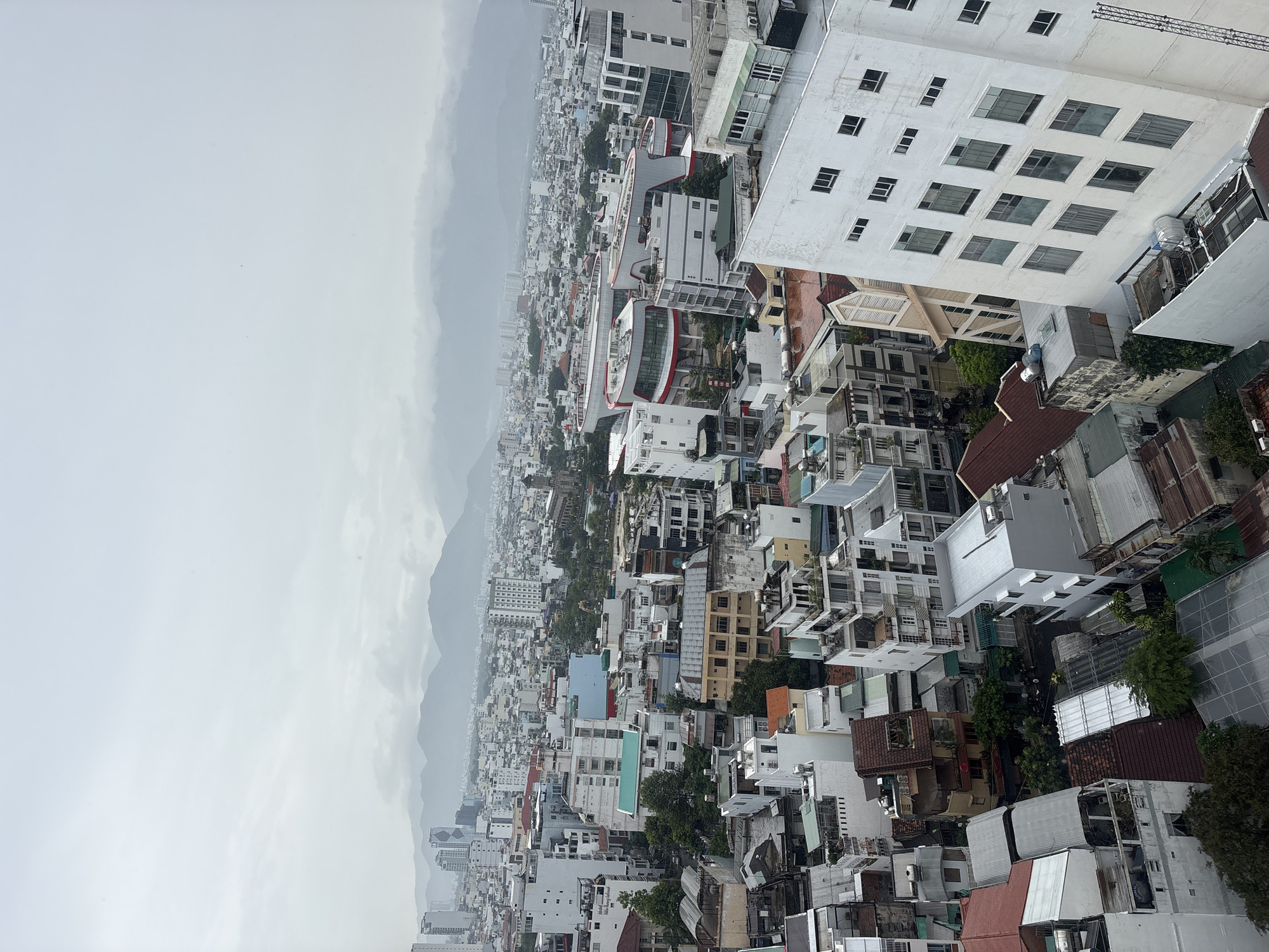The image presents an elevated view of Nha Trang Ward, Vietnam, depicting a dense urban landscape bordering mountainous terrain under overcast skies. The foreground is dominated by closely packed buildings varying in height and architectural style, predominantly white and beige, with some featuring brown tiled roofs. Visible amongst the structures are hints of greenery from trees interspersed within the urban fabric. The midground extends into a horizon line marked by indistinct grey mountains shrouded in mist, implying distance and creating a blurred backdrop to the city. The atmospheric conditions suggest either rain or heavy humidity, contributing to the muted color palette of the image.