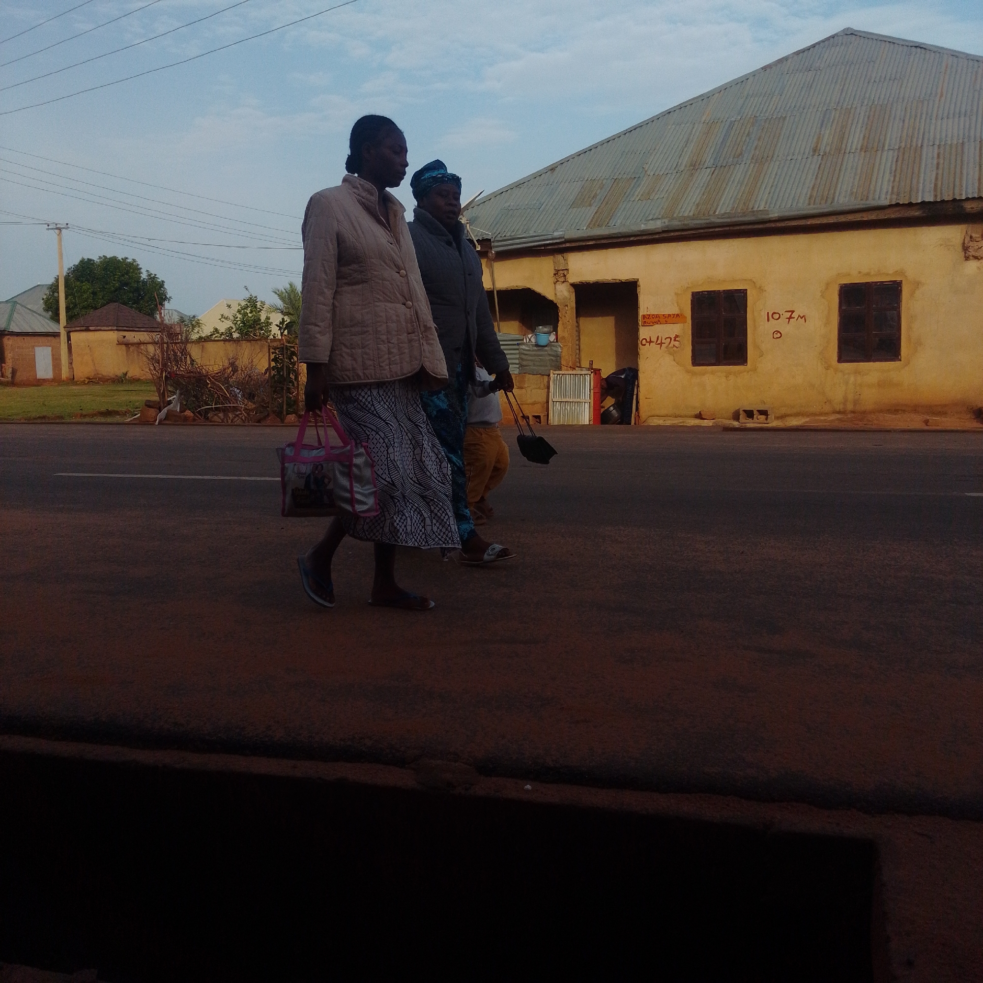 Two women walk on a paved road in Gombe, Nigeria. The woman on the left wears a light-colored quilted jacket and a patterned skirt. She carries a pink and silver patterned tote bag. The woman on the right wears a dark jacket and a blue and white headscarf. She pulls a black wheeled suitcase. A small child in orange pants walks between them, holding the suitcase handle. Behind them, a yellow building with a corrugated metal roof has two dark windows. Red graffiti is visible on the wall near the windows. A power line runs across the sky. The scene appears to be set during daylight.