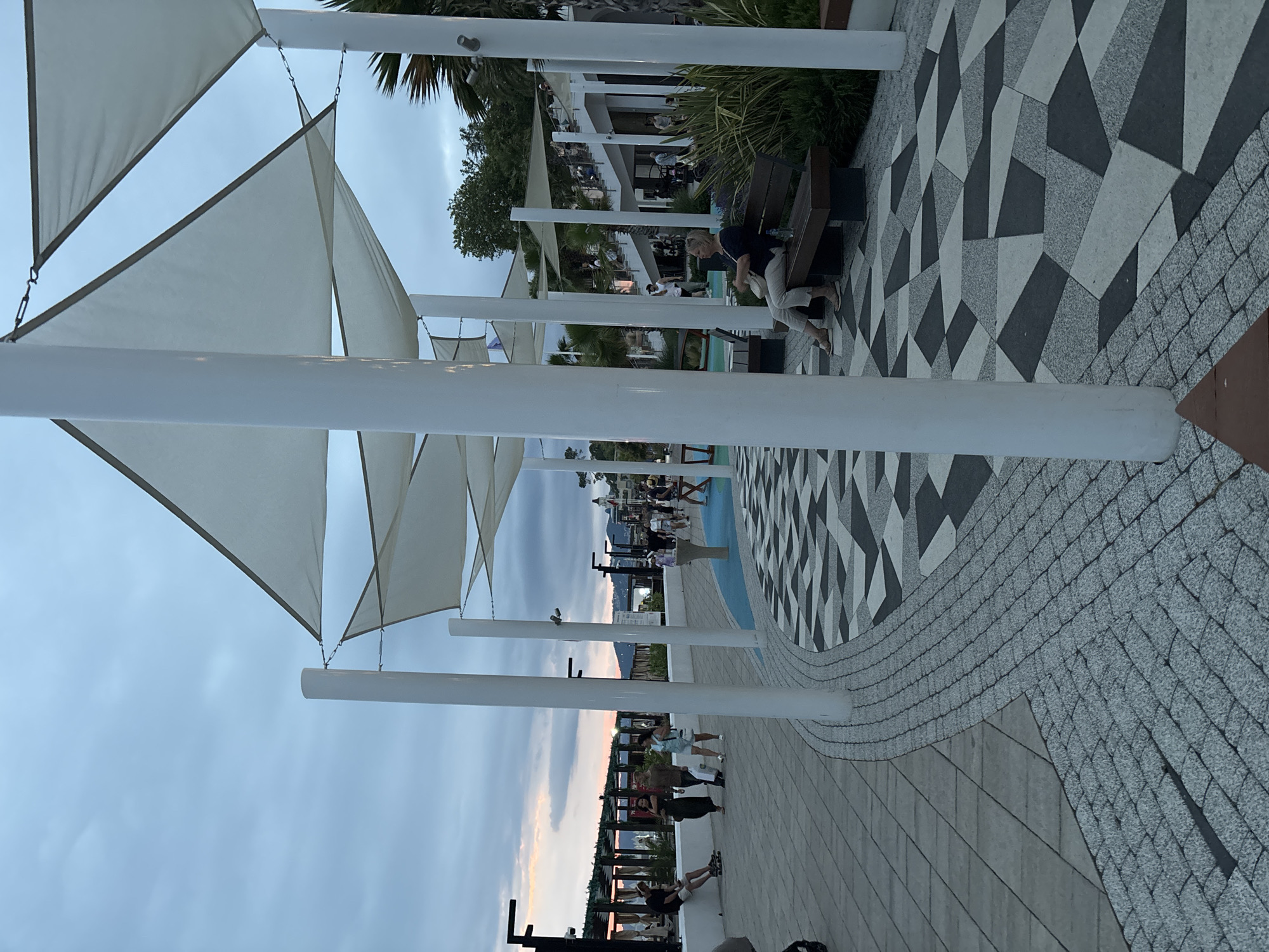 The image depicts a waterfront promenade in Sochi, Russia, at dusk. The sky is overcast with hints of sunset colors. Large, white fabric sails are suspended overhead, supported by white poles, providing shade. The ground is paved with a patterned arrangement of light and dark grey tiles, with a cobblestone border.
Several people are visible on the promenade. On the left, a man sits on a low wall on rollerblades. Further down, a group of people walks along the promenade. In the foreground on the right, a woman sits on a dark brown bench, facing left, holding a light-colored bag. Behind her, lush green foliage and palm trees are visible. In the background, a multi-level building with stairs is illuminated, and more people are seen walking. Lights line the promenade, and structures resembling covered seating areas are present along the edge of the water. The overall scene conveys a relaxed, evening atmosphere on a public gathering space.